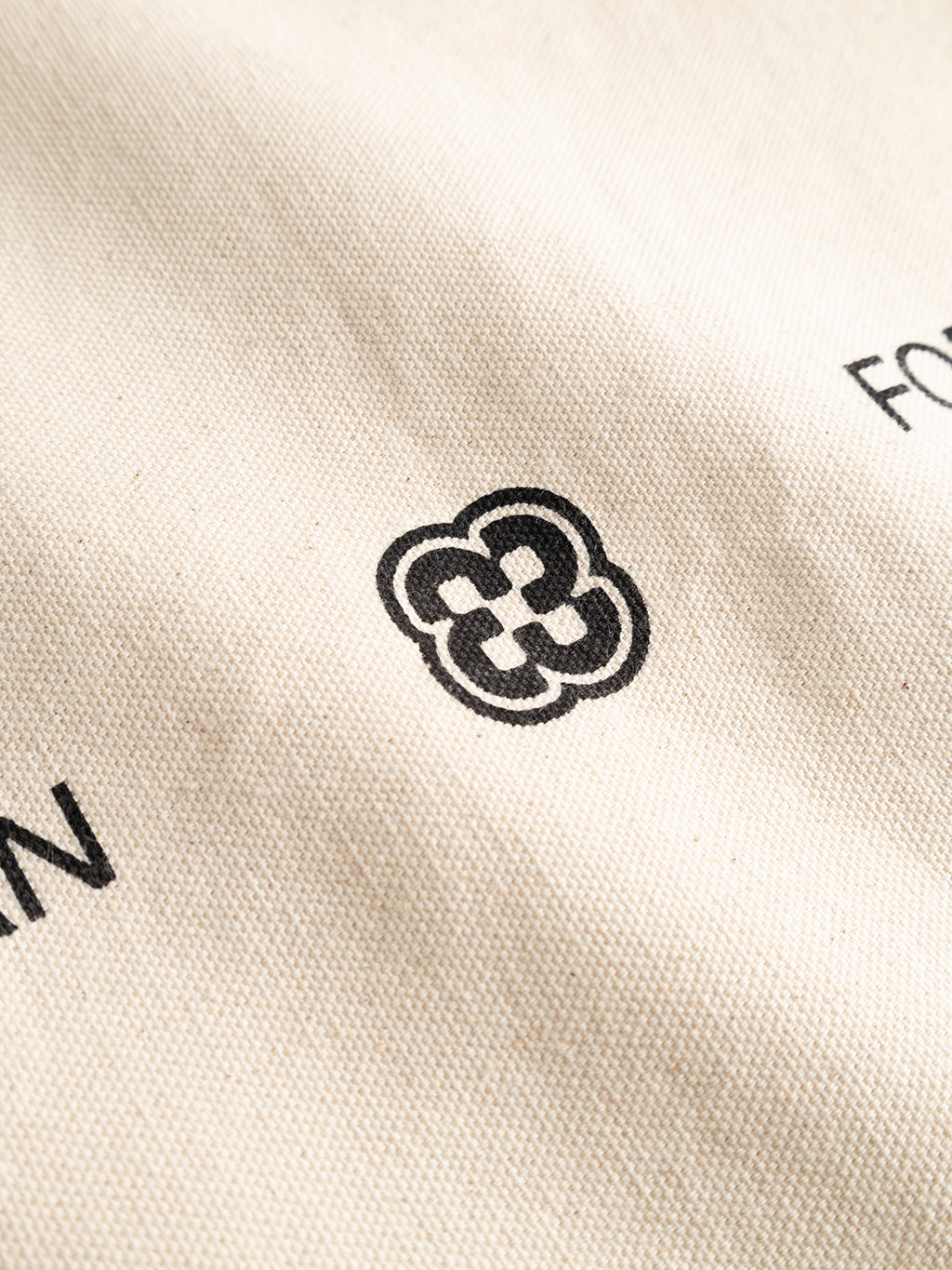 Close-up of an off-white GABworn Tote Bag featuring a black, four-leaf clover-style Gabba emblem and partial black lettering.