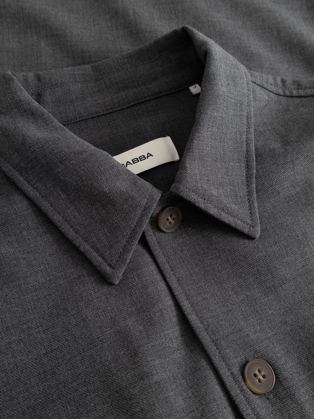 Close-up of the GABtogger Push Phin LS Overshirt GRS in dark grey, featuring a classic collar and brown buttons. A white tag inside the collar displays the Gabba brand name. Made from a soft, textured wool-viscose blend, this regular fit overshirt is a versatile layering essential for any wardrobe.