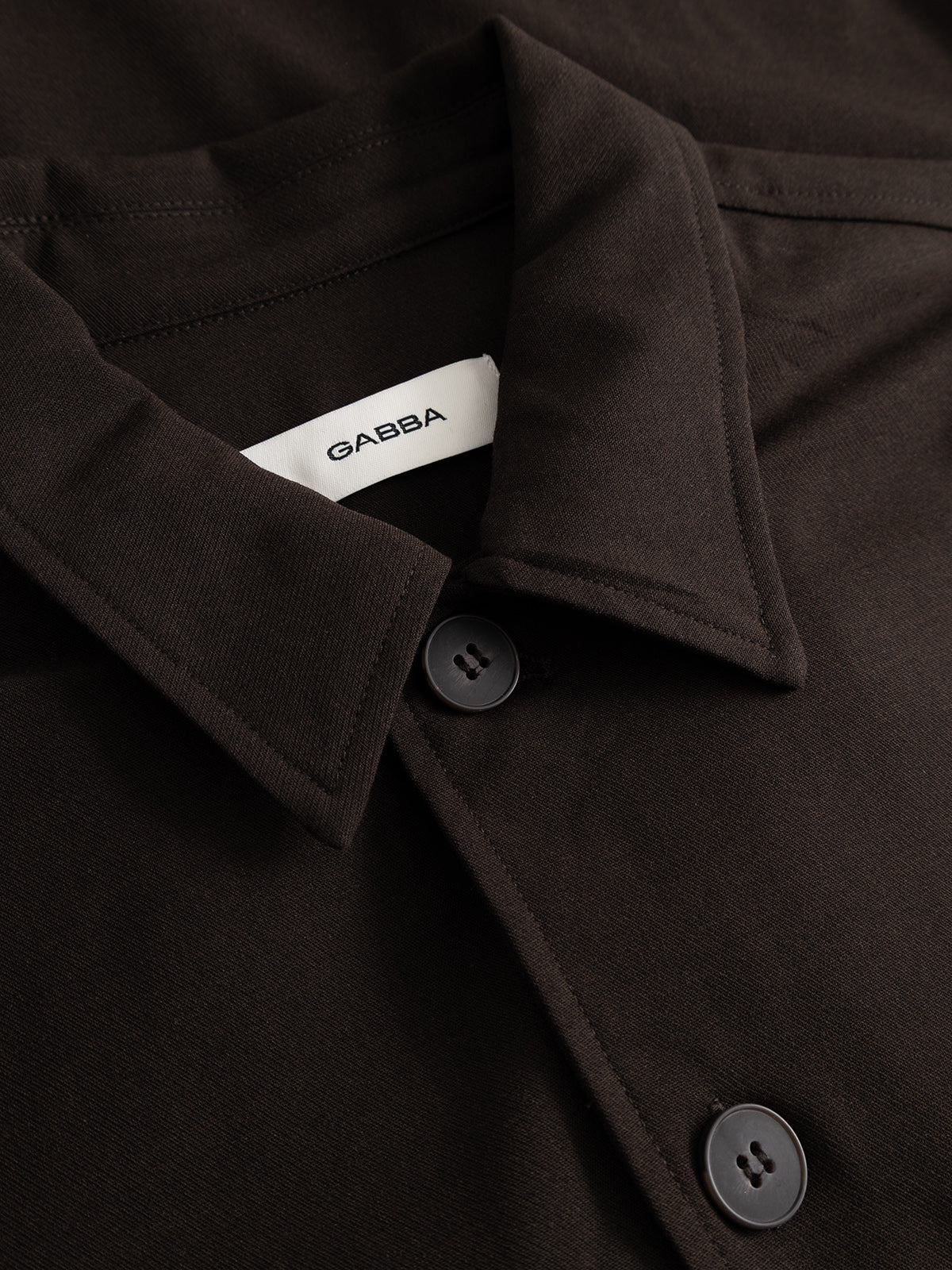 Close-up of the GABtogger Push Phin LS Overshirt GRS in Brown Slate by Gabba, featuring a dark brown collar with two visible buttons and a white Gabba tag inside. Crafted from a soft, smooth wool-viscose blend, this versatile overshirt offers both comfort and style—making it an essential layering piece.