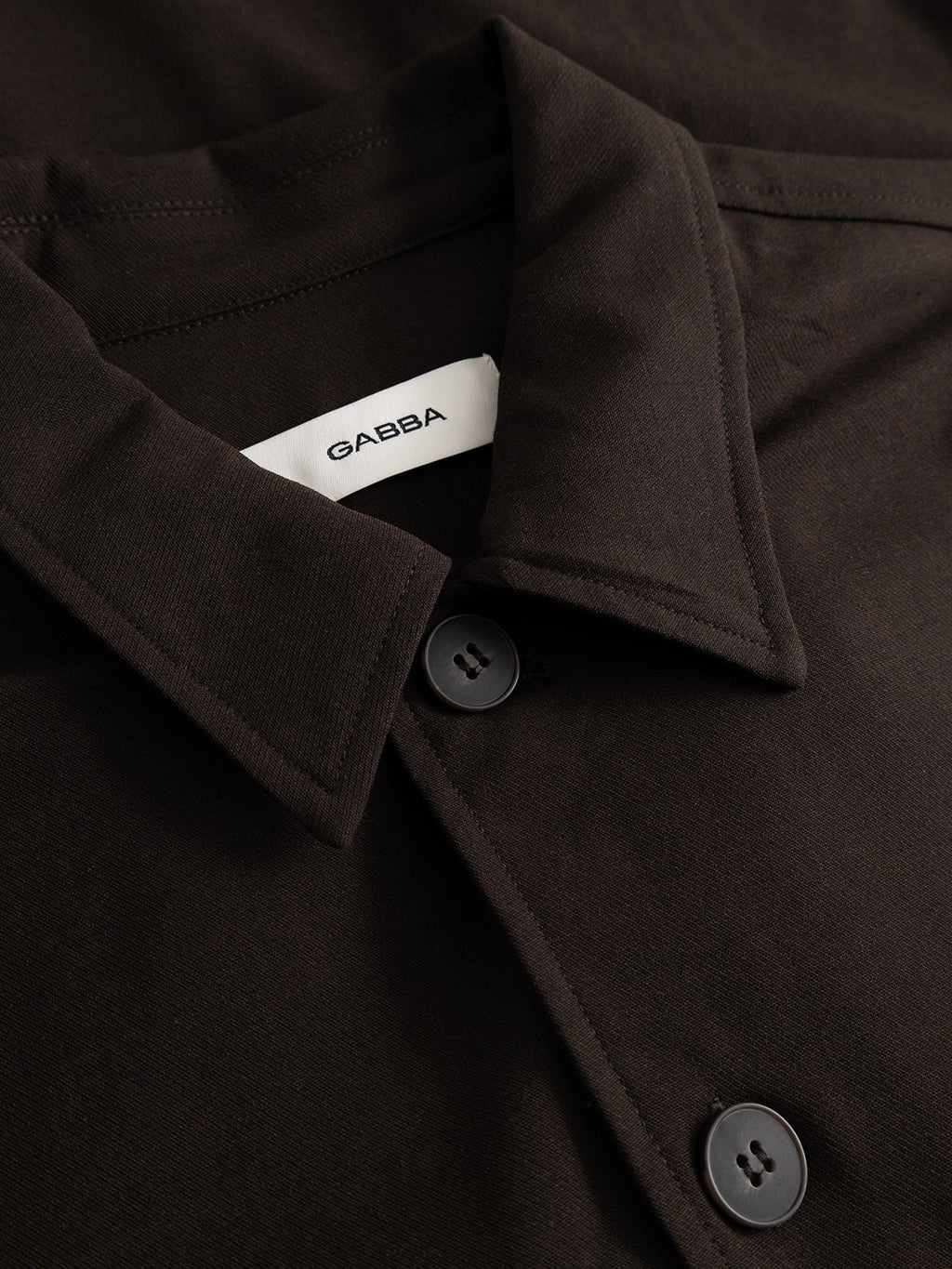 Close-up of the GABtogger Push Phin LS Overshirt GRS in Brown Slate by Gabba, featuring a dark brown collar with two visible buttons and a white Gabba tag inside. Crafted from a soft, smooth wool-viscose blend, this versatile overshirt offers both comfort and style—making it an essential layering piece.