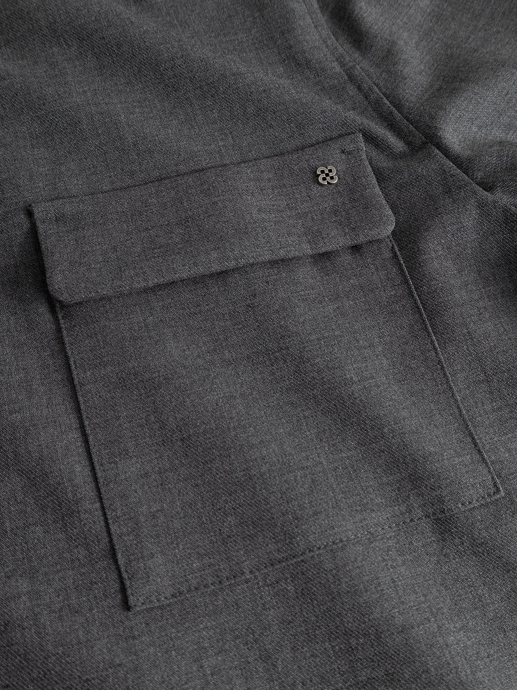 A close-up of the GABtogger Push Phin LS Overshirt GRS in Dk. Grey by Gabba reveals a gray fabric pocket with a flap, accented by a small decorative metal emblem in the top right corner. Expertly crafted from a premium wool-viscose blend, this layering essential by Gabba embodies durability and timeless style.