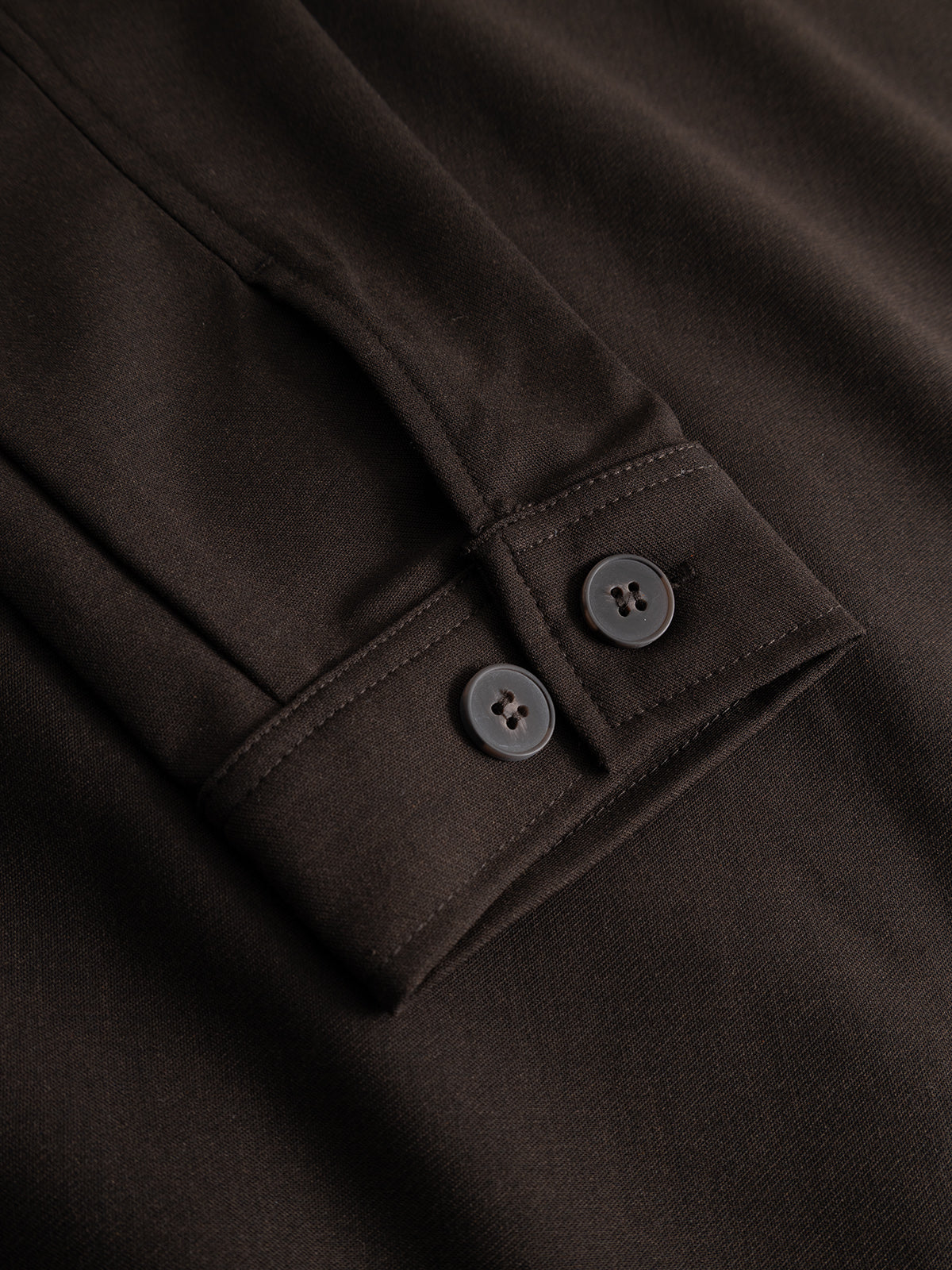Close-up of the Gabba GABtogger Push Phin LS Overshirt GRS in Brown Slate, showcasing a dark brown cuff with two black buttons and visible stitching. Crafted from a soft wool-viscose blend, this regular fit overshirt is displayed against a matching fabric background, making it an ideal layering essential for any wardrobe.