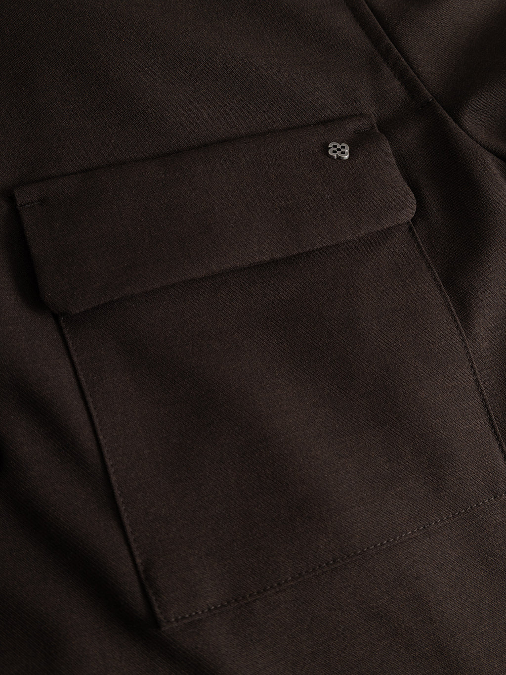 Close-up of the GABtogger Push Phin LS Overshirt GRS by Gabba in Brown Slate, showcasing a dark brown wool-viscose blend fabric pocket with a flap closure. A small metallic emblem is attached to the top corner of the flap, which features a concealed snap-button closure. The texture of the fabric is clearly visible.