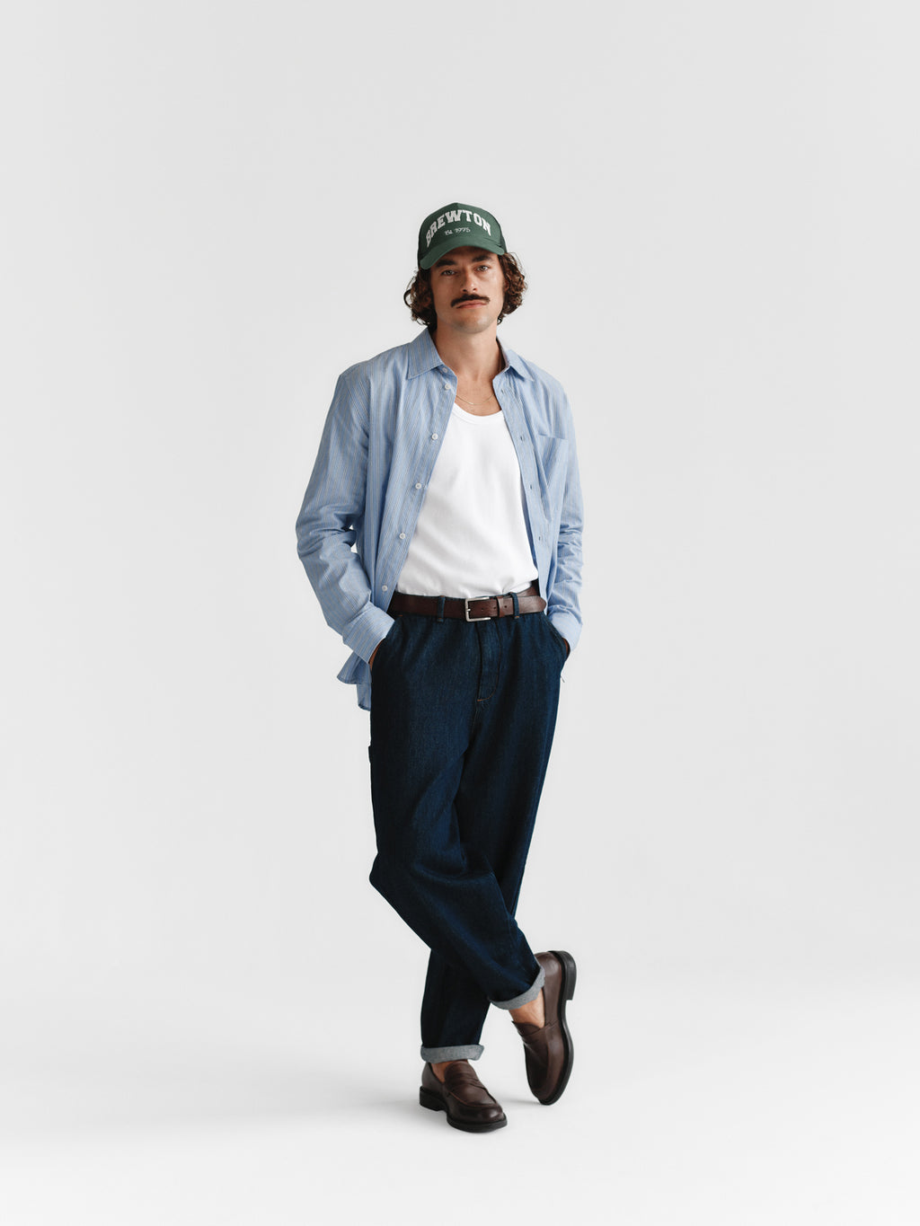 A man with a mustache, green cap, light blue shirt, white tank top, Gabba Vito - Dark Blue Denim pants, and brown loafers stands with hands in pockets against a plain white background.
