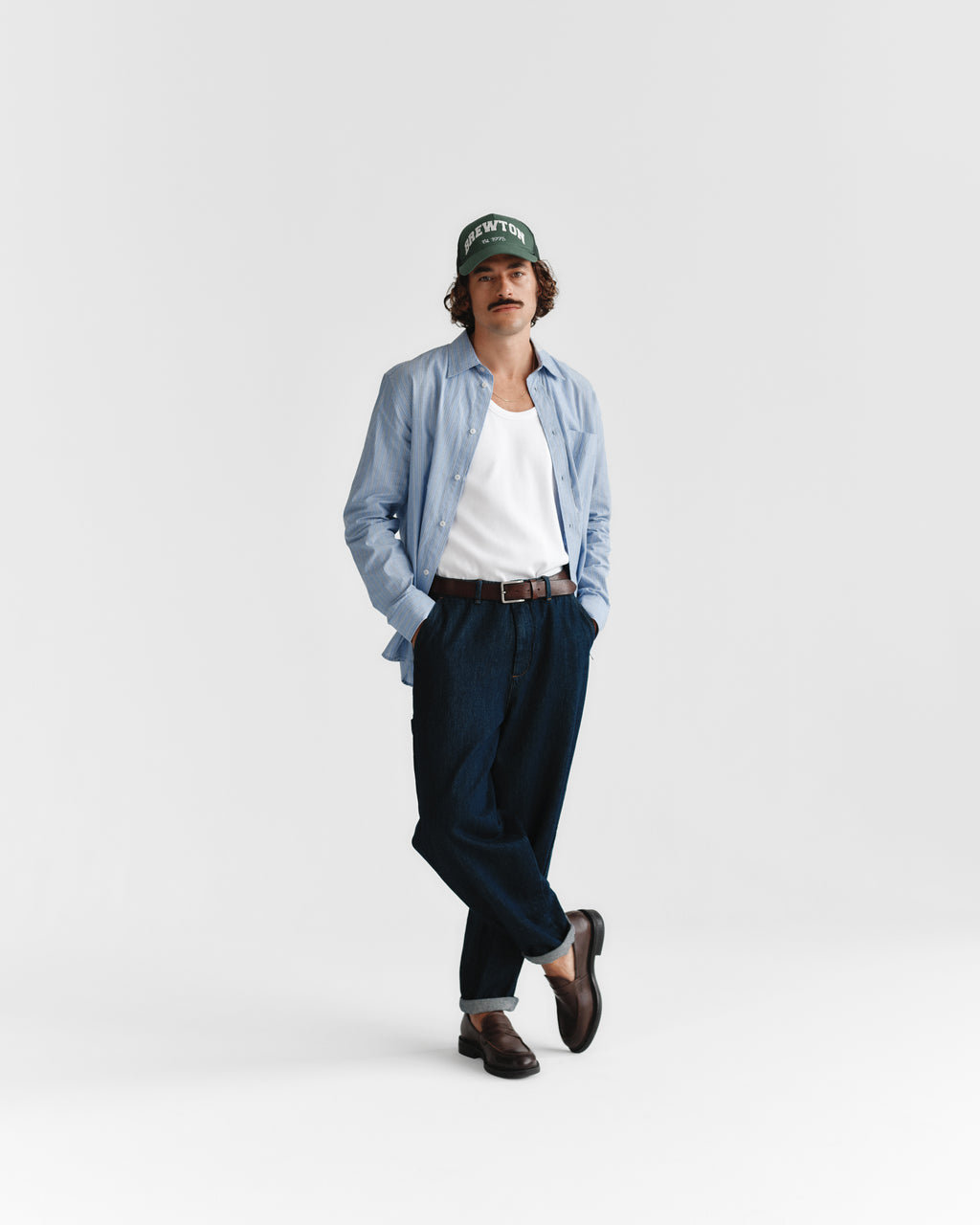 A man in a green beanie, light blue button-up over a white tank, dark pants, and brown loafers stands against a white backdrop. He finishes his look with the Gabba Stitch belt - Cognac, featuring subtle stitch details.