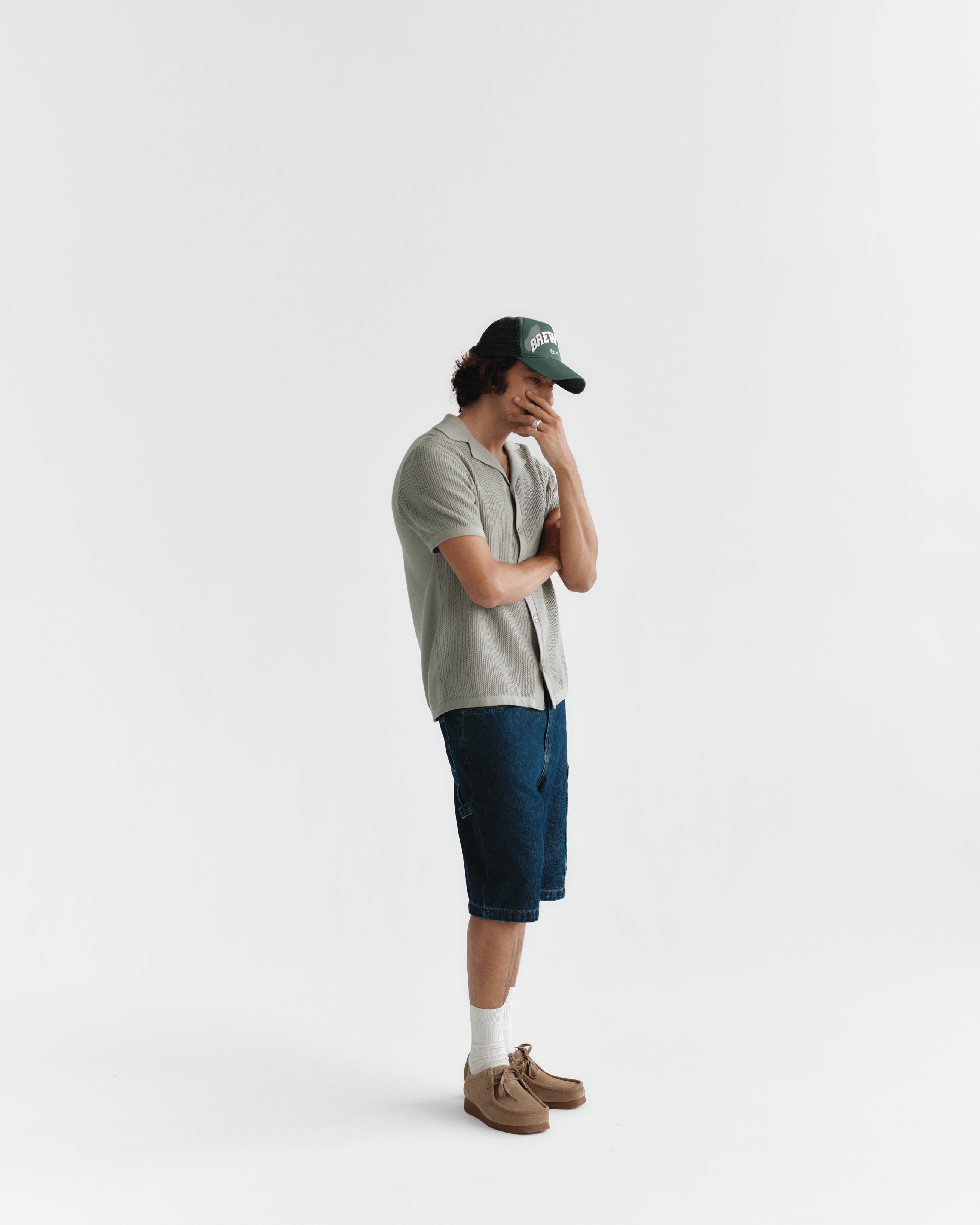 A person is standing against a white background wearing a green cap, light gray short-sleeve shirt, and Ugo Cargo Fone - Mid Blue Denim by Gabba. The blue shorts have a loose fit, paired with white socks and brown shoes. They look down with one hand covering part of their face.