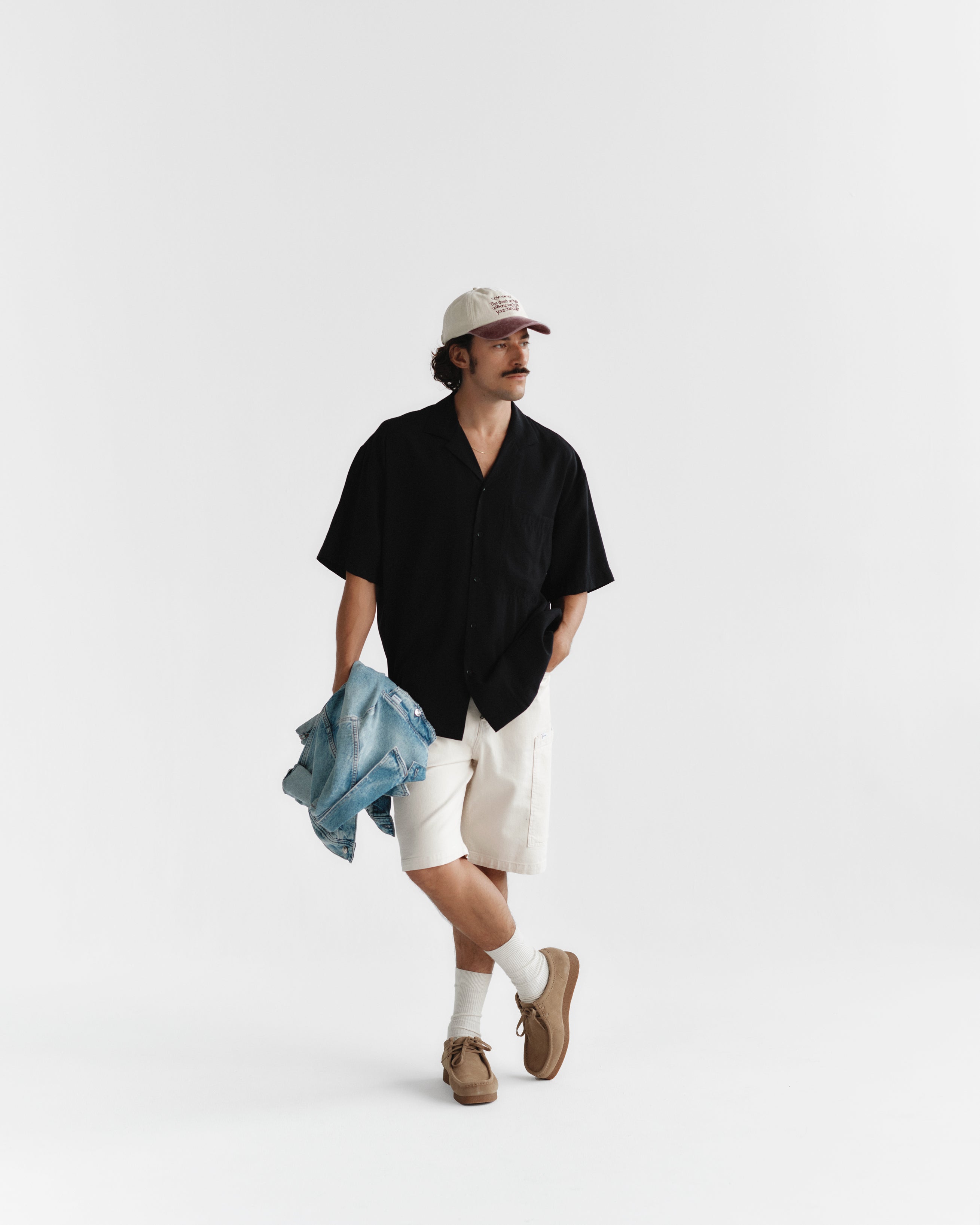 A mustached man wears a Gabba Jens Over Resort Yari SS - Meteorite shirt, beige shorts, and brown shoes while holding a denim jacket. He stands against a plain white background, looking to the side with one hand in his statement chest pocket and sports a beige baseball cap.