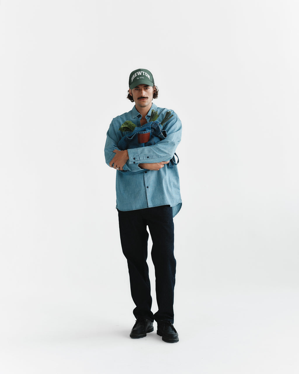 A person wearing the Gabba Jens Over Topaz LS in light chambray and black pants stands holding a potted plant against a plain white background.