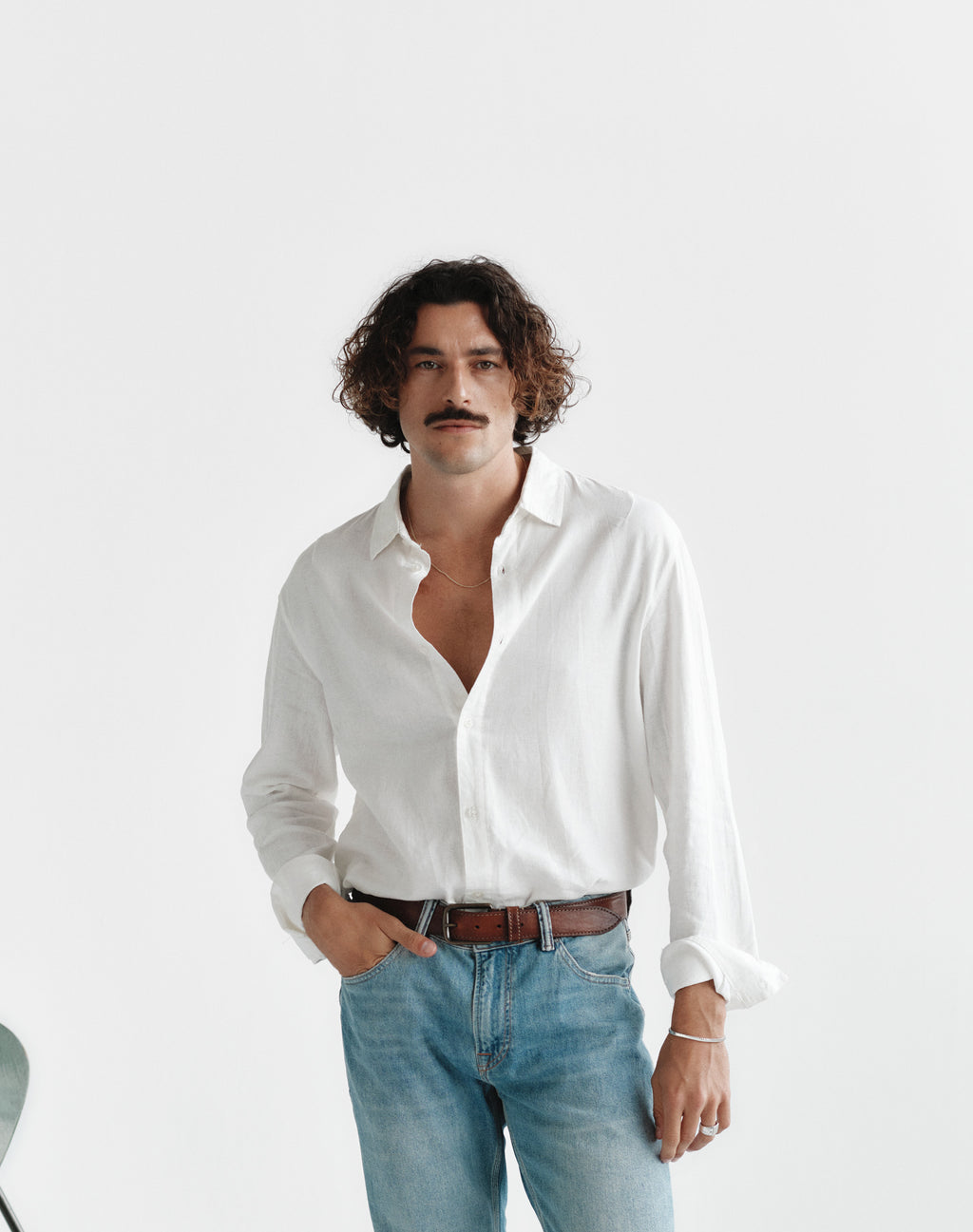 A man with curly hair and a mustache stands against a plain white background, wearing the Gabba Sven Reg Lino LS - Off White linen button-up shirt and light blue jeans, with one hand in his pocket.