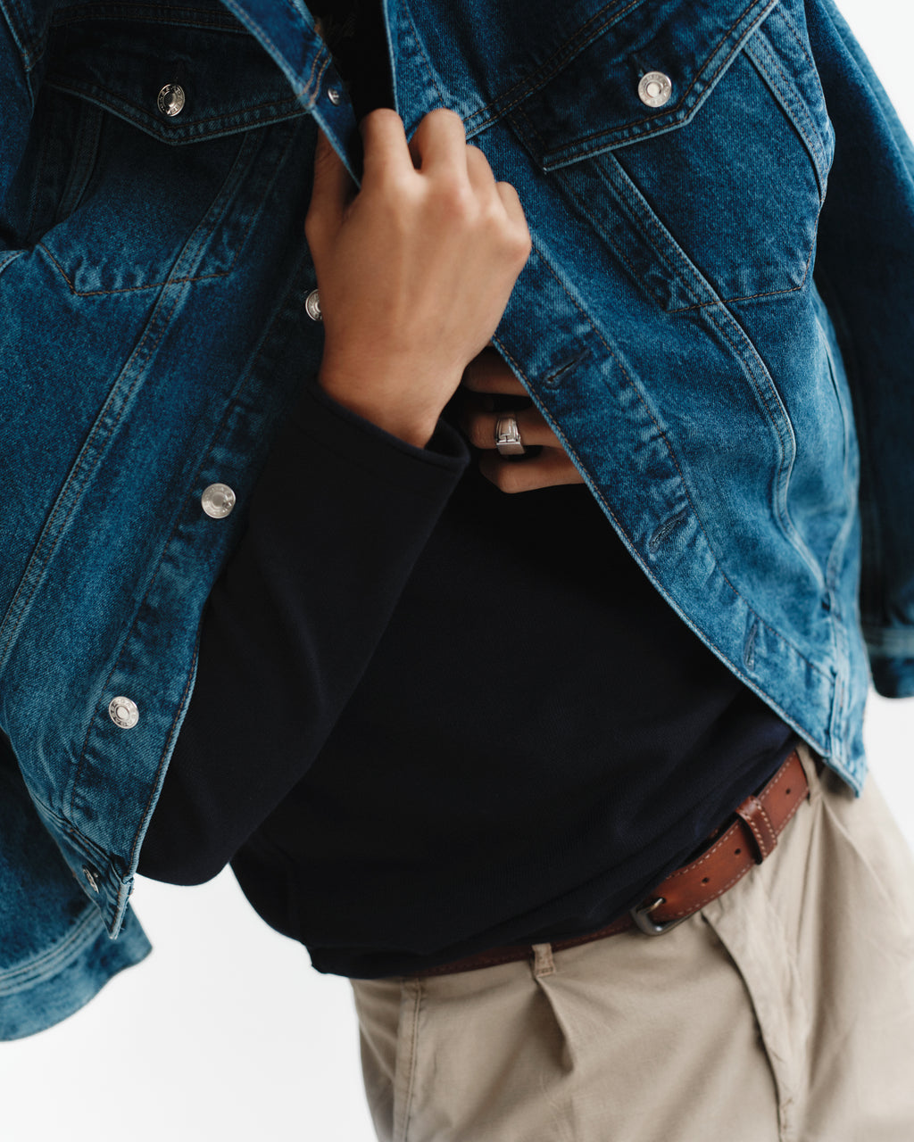 A person wears a blue denim jacket, black shirt, and beige pants accessorized with the Gabba Stitch belt in Cognac, holding the jacket open to reveal a silver ring. The image is cropped to highlight the torso.