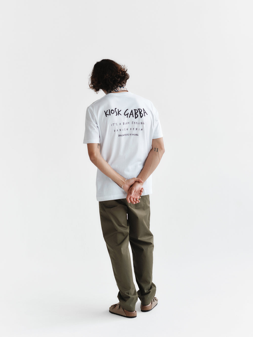 A person with curly hair stands facing away, hands behind back, wearing the Gabba Dune Kiosk B SS GOTS - White premium organic cotton T-shirt with black text, olive green pants, and sandals against a plain white background.