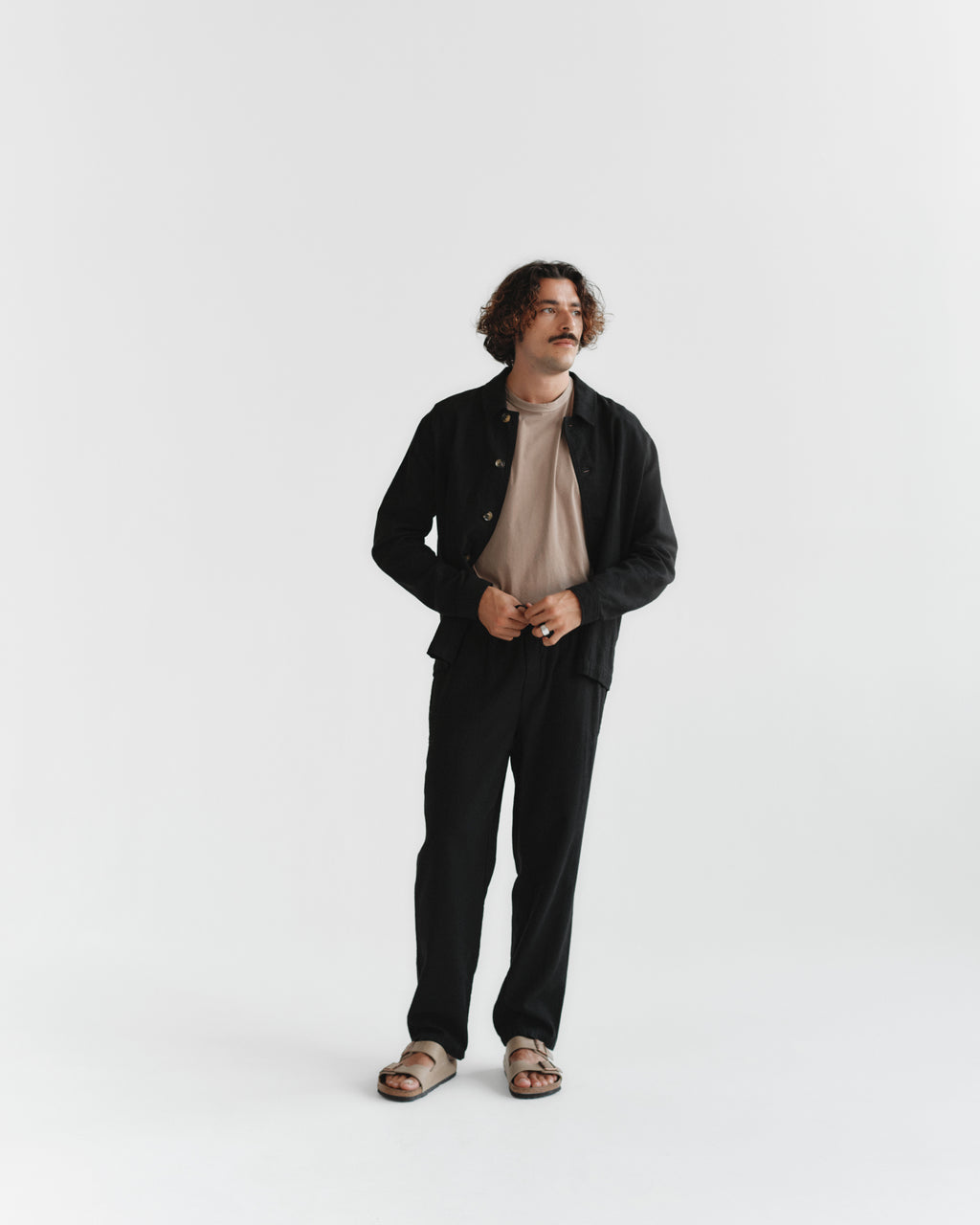 A man with curly hair and a mustache stands against a white background, wearing the Gabba Lagos SS - Safari t-shirt under a black jacket, paired with classic fit black pants and brown sandals. He gazes to the side, hands clasped.