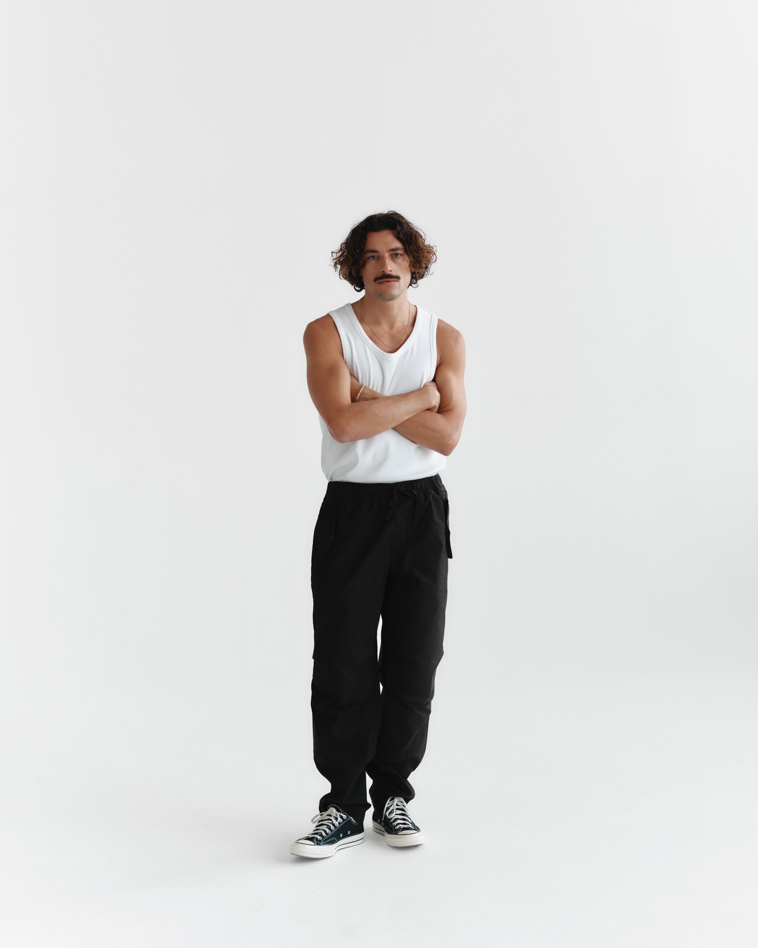 A person with curly hair and a mustache stands against a plain white background, wearing a white Orsen tank top by Gabba, black pants, and black sneakers. Their arms are crossed in a relaxed pose, embodying the essence of easygoing summer days.