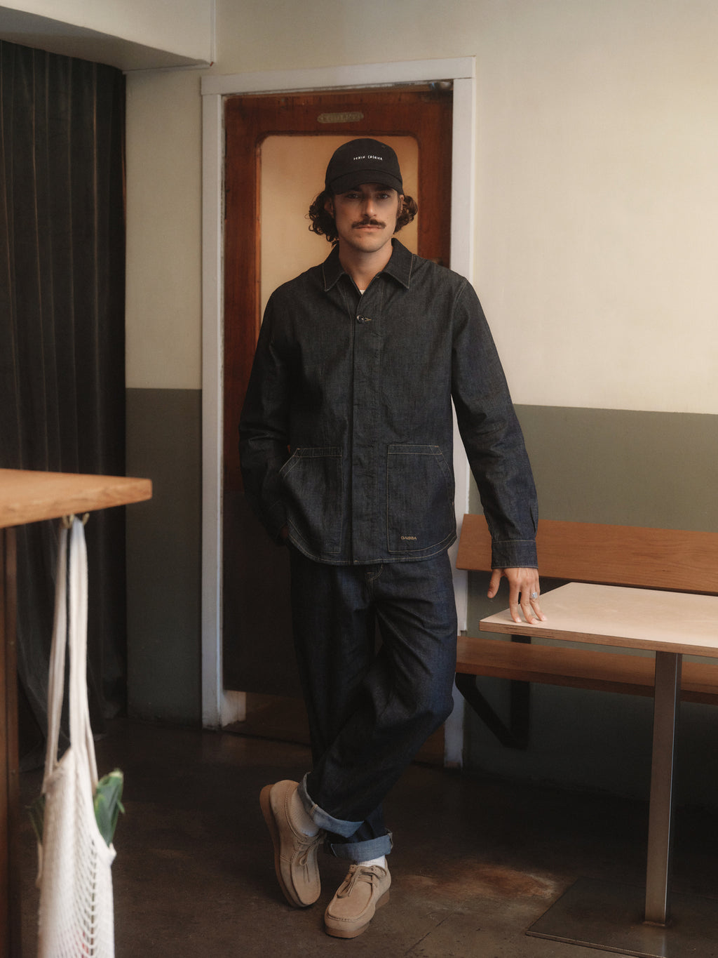 A man with a mustache wears Gabbas Jap Beam - Dark Blue Denim jacket and pants made from 100% cotton selvage, pairing them with beige shoes and a black cap as he leans casually on a wooden table in a minimalist room.