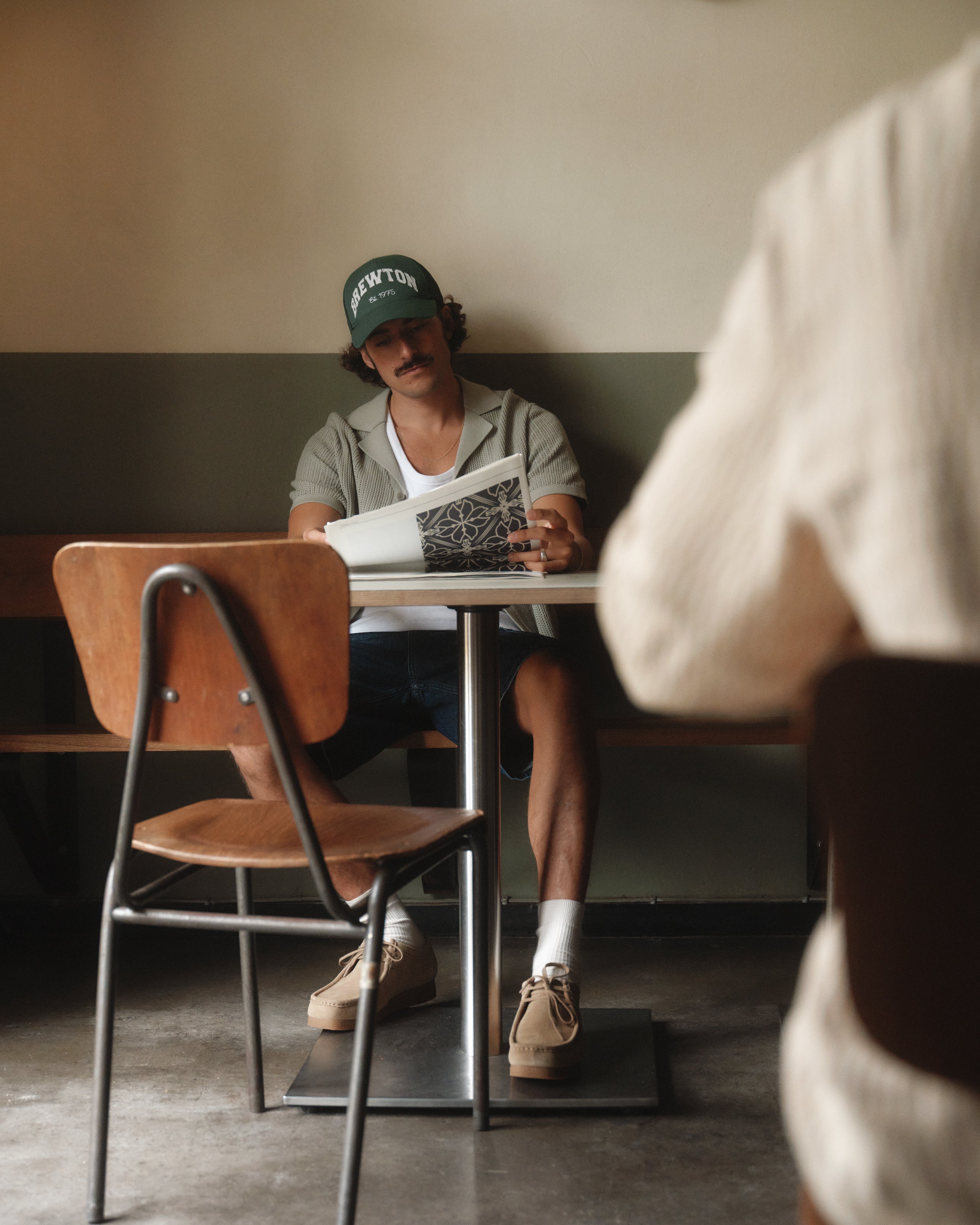 Someone wearing the Gabba Geno Resort SS in Light Green and casual attire, along with a premium knit cotton cardigan, sits at a cafeteria table flipping through a magazine. An empty chair is in the foreground with another seated person blurred. The rooms calm, retro vibe features a neutral color palette.