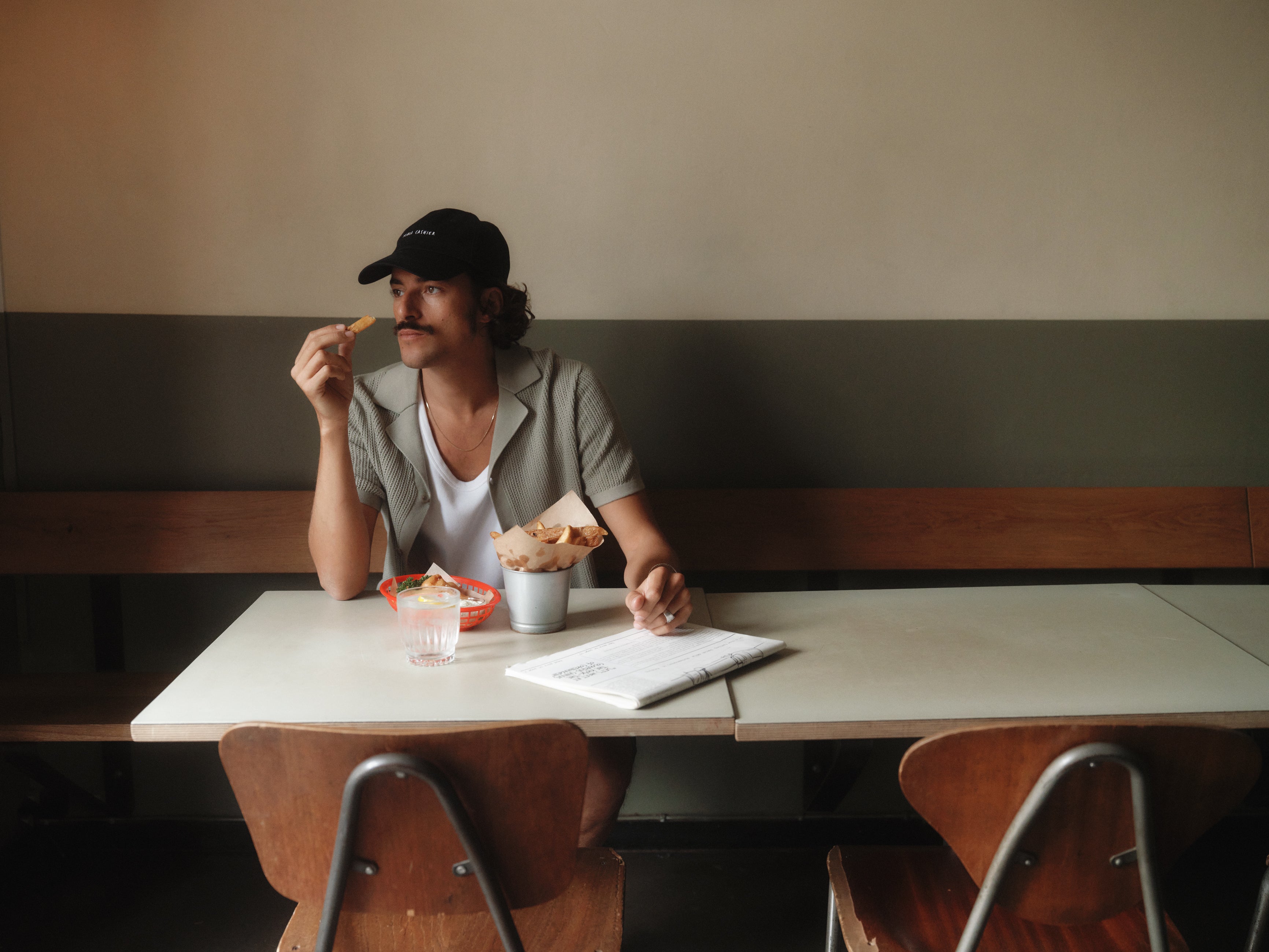 A person with a mustache sits at a table, wearing a cap and Gabbas Geno Resort SS - Light Green shirt. They enjoy a pastry from a bowl, with a drink and magazine on the table. Two empty chairs are in the foreground. The setting looks casual and relaxed.