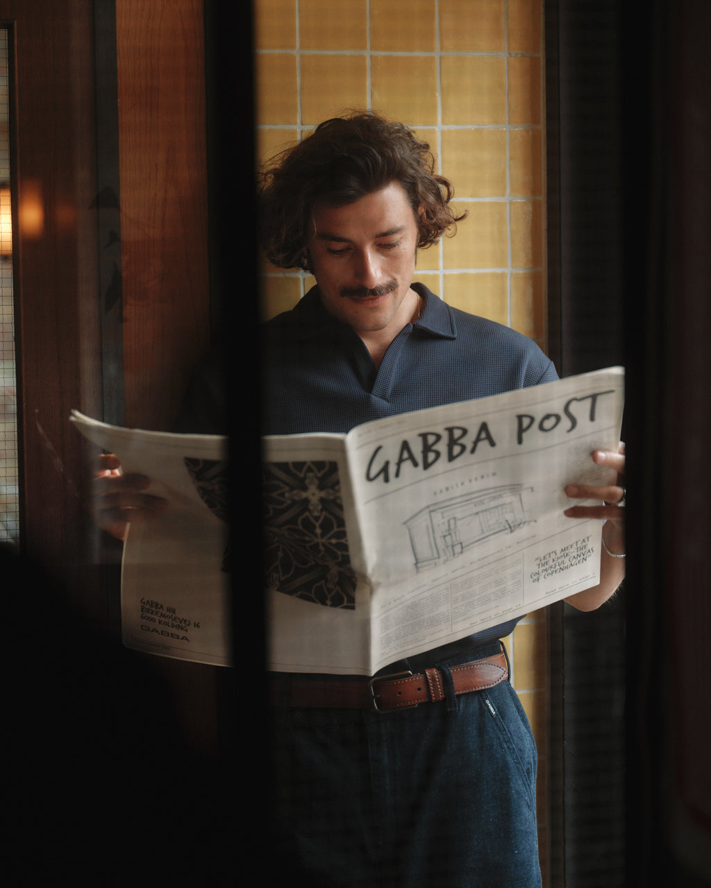 A man with wavy hair and a mustache reads the GABBA POST indoors near yellow tiles, wearing a dark shirt, blue trousers, and the Gabba Stitch belt in Cognac—a leather belt with subtle stitch details.