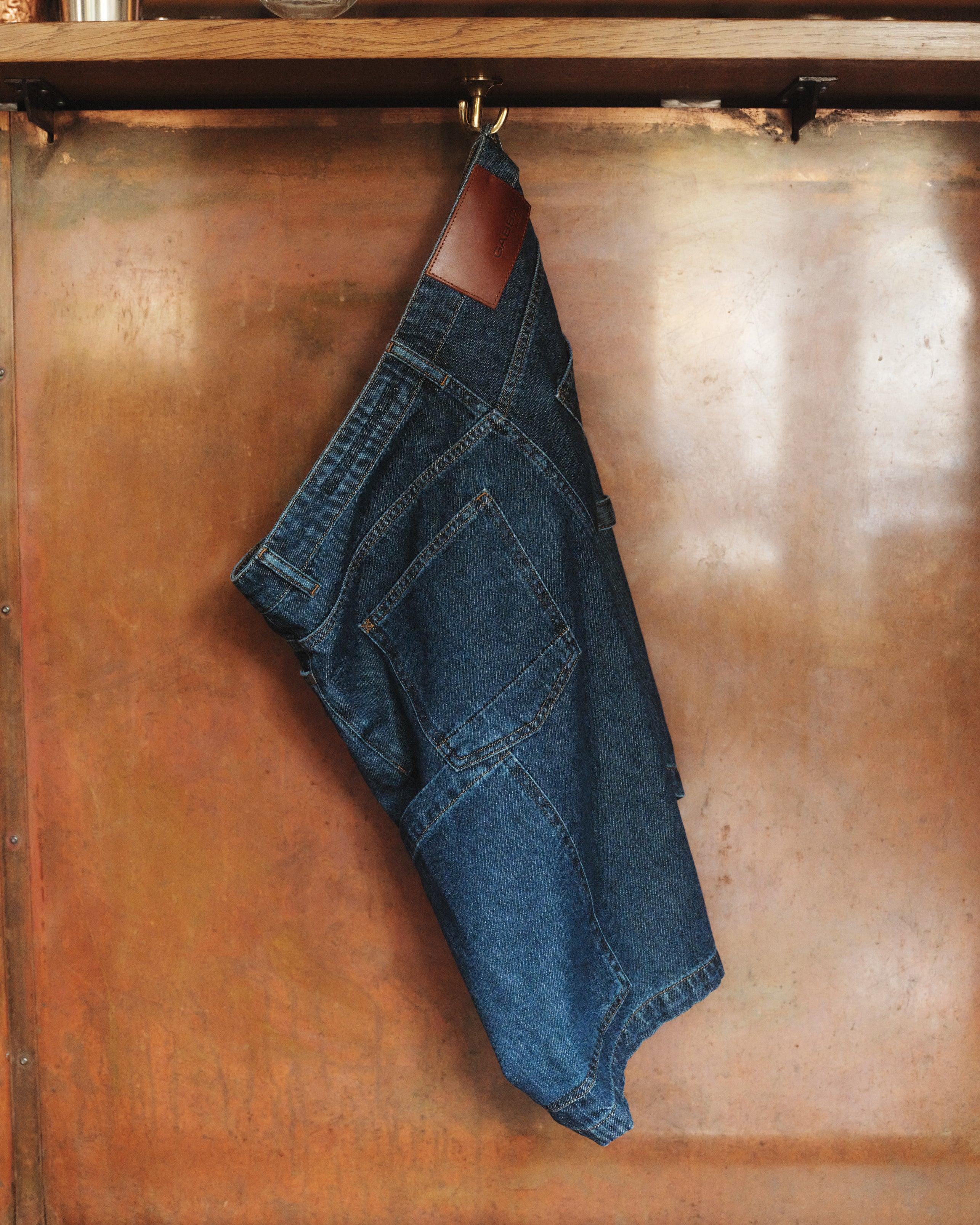 The Ugo Cargo Fone - Mid Blue Denim by Gabba, with its classic workwear style, hangs on a hook against a copper backdrop. Its loose fit reveals a visible back pocket and leather patch on the waistband, merging timeless style with functionality.
