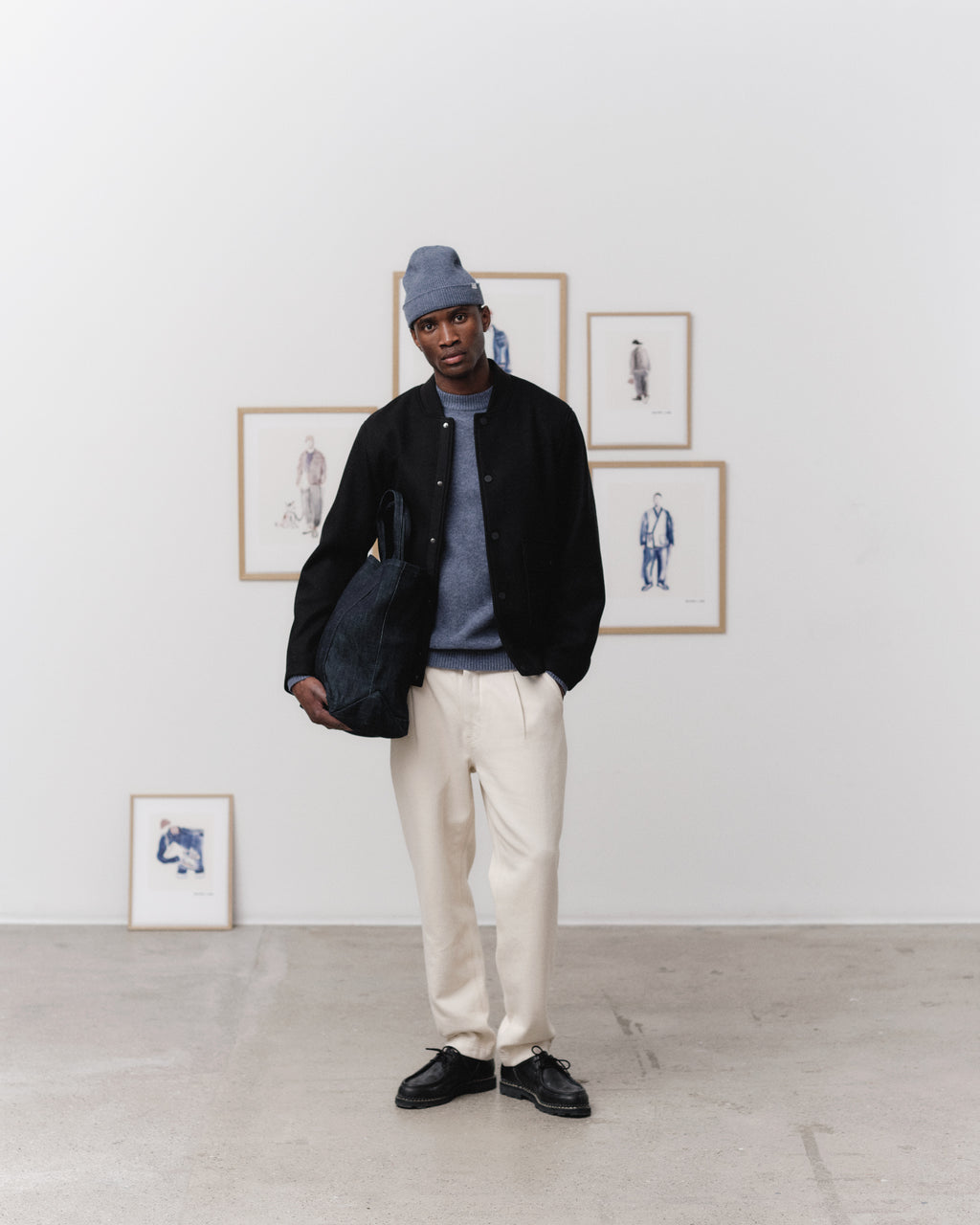 A man stands indoors by framed fashion sketches, wearing a gray beanie, dark jacket, blue sweater, Gabba Joe-Pas Vill Lt. Sand relaxed-fit cotton twill trousers with black shoes, holding a dark bag and one hand in his pocket.