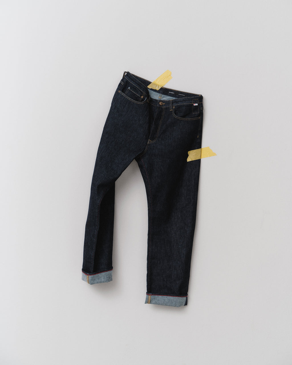 The Noah Self - Dark Blue Denim jeans by Gabba, featuring a loose straight fit and rolled cuffs, made from recycled fibers, are taped to a plain white wall with two pieces of yellow tape.