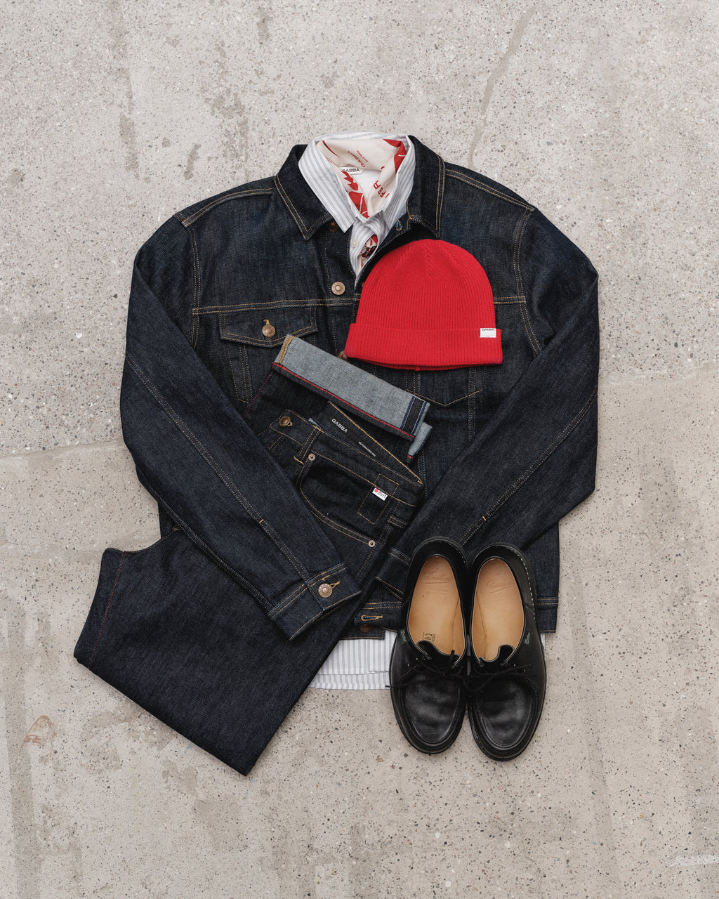 A flat lay displays Gabba’s Luca Self Dark Blue Denim jeans and jacket, a white shirt with a red pattern, a red beanie, and black dress shoes on a light textured surface.