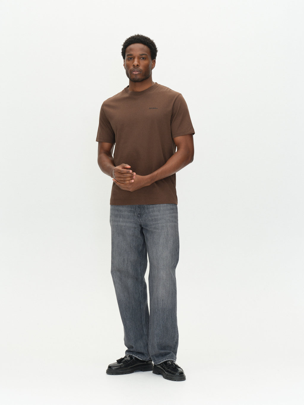 A man wearing a brown GABdune Logo GOTS SS T-Shirt with a small logo on the chest, grey jeans, and black loafers.