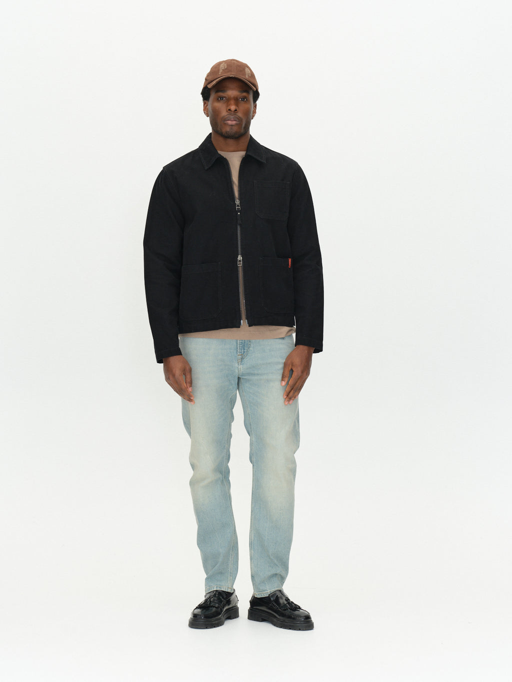 A man wears a black zip-up jacket, tan t-shirt, brown cap, and GABcarl CrosV2 Jeans - Lt. Blue Denim in a loose tapered fit.