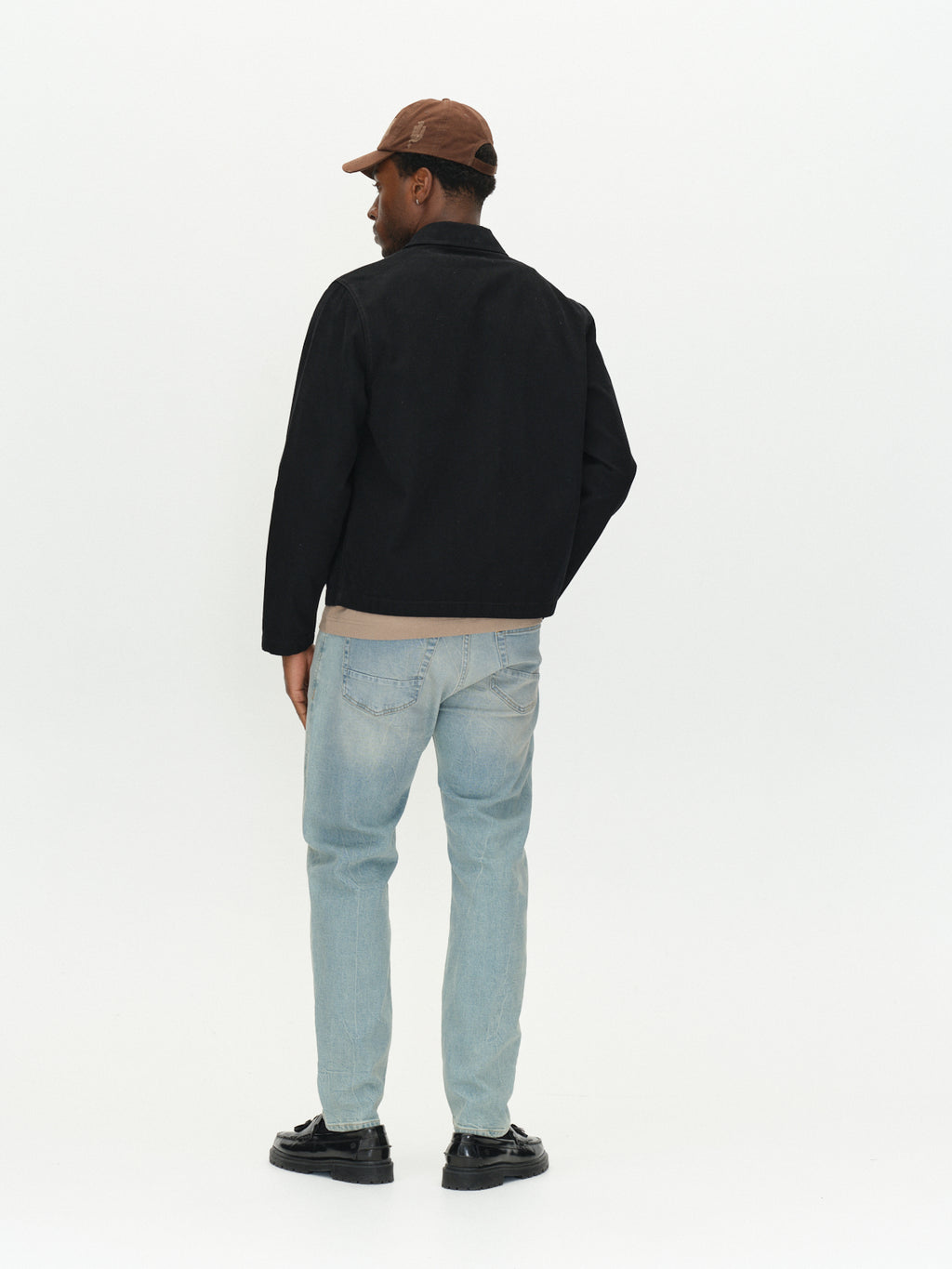 A person seen from behind, wearing a brown baseball cap, a black jacket, GABcarl CrosV2 Jeans - Lt. Blue Denim, and black loafers.