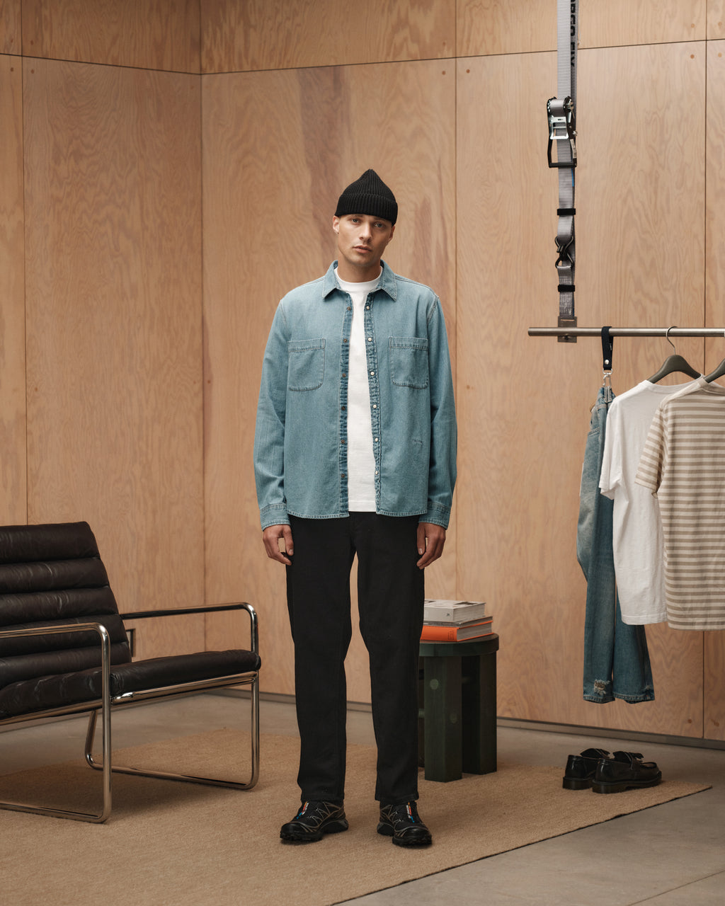 A man wears Gabbas Max Sword - Black Denim pants, paired with a black beanie, light denim shirt, and white t-shirt, standing in a stylish room with wood-paneled walls, black chair, hanging clothes, books, and a carpeted floor.
