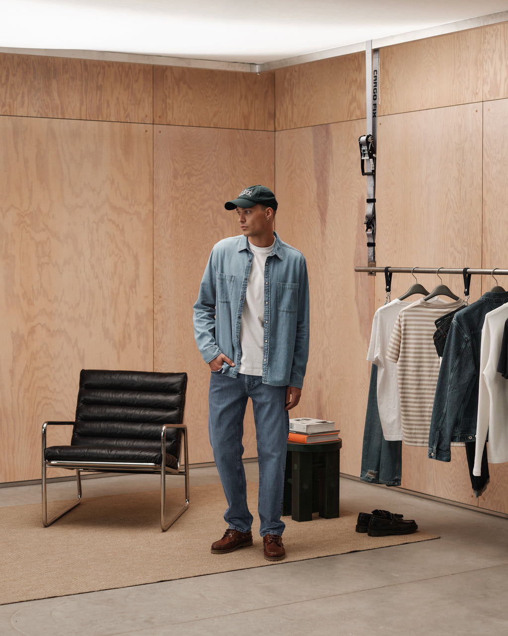 Wearing Gabbas Marc F1010 - Dark Blue Denim jeans, a person in a denim shirt, white tee, and green cap stands in a modern minimalist room with wood-paneled walls, a black chair, hanging clothes, and carpeted floor for a classic look.