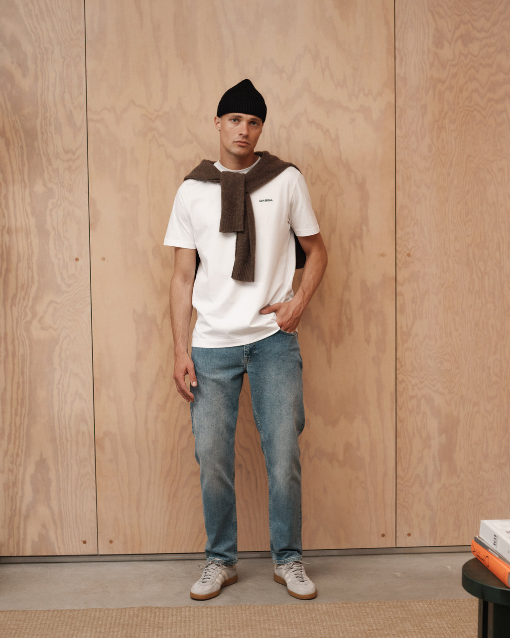 A man with a modern look stands indoors against a light wooden wall, wearing Gabba Marc 5poc Mid Blue Denim jeans, a black beanie, white t-shirt, beige sneakers, and a brown sweater draped over his shoulders.