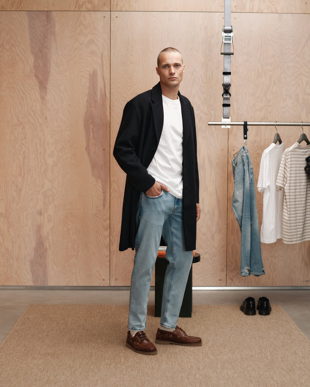 A man stands indoors in a black coat, white shirt, and regular fit jeans for a modern classic look. Behind him, a rack displays the Gabba Marc F1012 - Lt. Blue Denim jeans, a striped t-shirt, and shoes against a wooden wall.