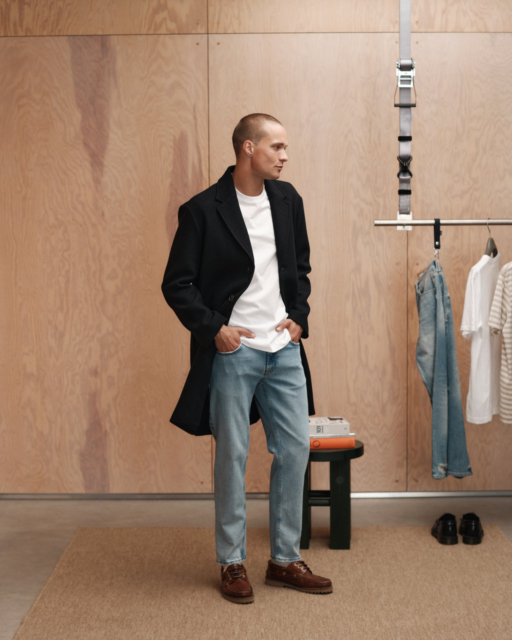 A man in a black coat, white shirt, and Gabba Marc F1012 - Lt. Blue Denim jeans stands indoors with hands in his pockets. Behind him are a wooden wall, hanging jeans, a striped shirt, shoes, and a small table with books.