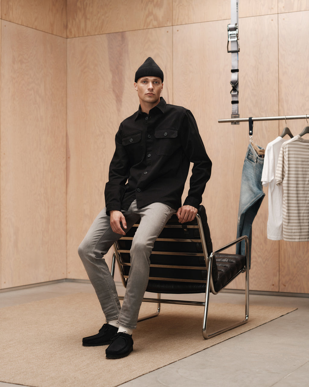 A man in slim fit gray pants, a black beanie, and jacket sits on a modern chair in a minimalist room. Behind him, a rack displays GABBA Rey K3454 - Denim wash jeans and striped shirts for a laid-back vibe.