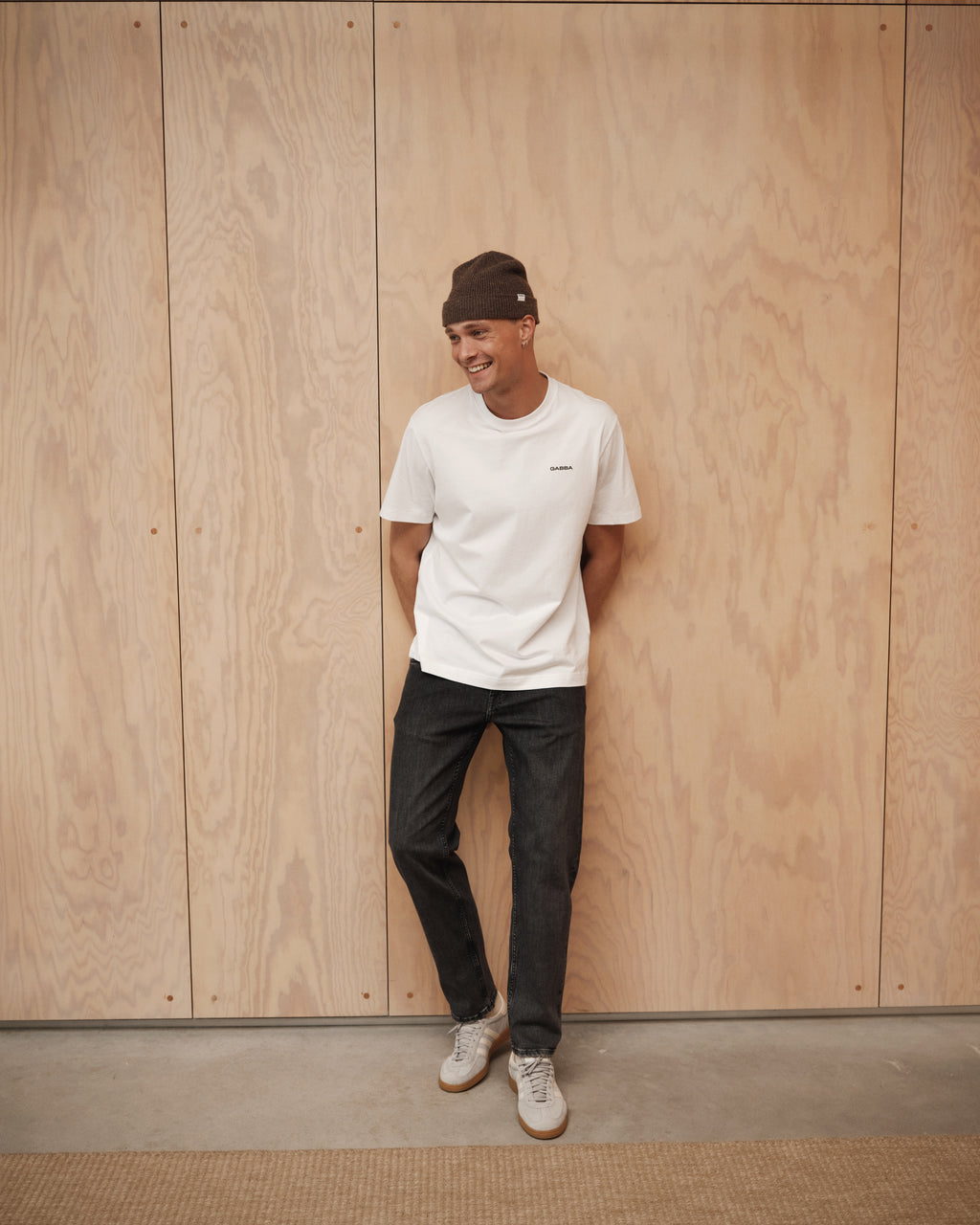 A man in a brown beanie and white t-shirt smiles with his hands in his pockets, wearing Gabba Marc F1011 Black Denim jeans and leaning against a light wood-paneled wall for a modern classic look.