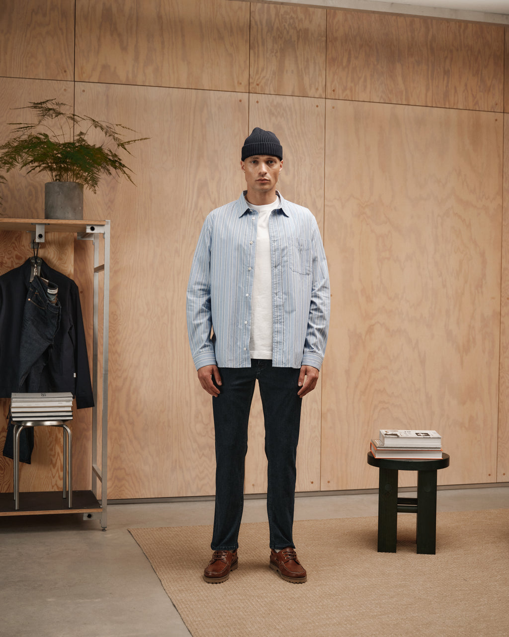 A man stands indoors on a beige rug, wearing Gabbas Marc Panter - Dark Blue Denim, a blue beanie, light blue striped shirt over a white tee, dark pants, and brown shoes. Nearby are a metal clothes rack and a small table with books.