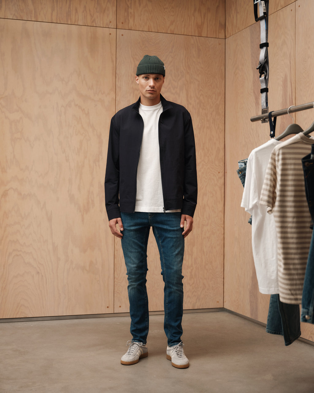 A man wearing a green beanie, black jacket, white shirt, beige sneakers, and slim Gabba Jones K3870 - Mid Blue Denim jeans stands in a wooden fitting room with hanging clothes visible to his right.