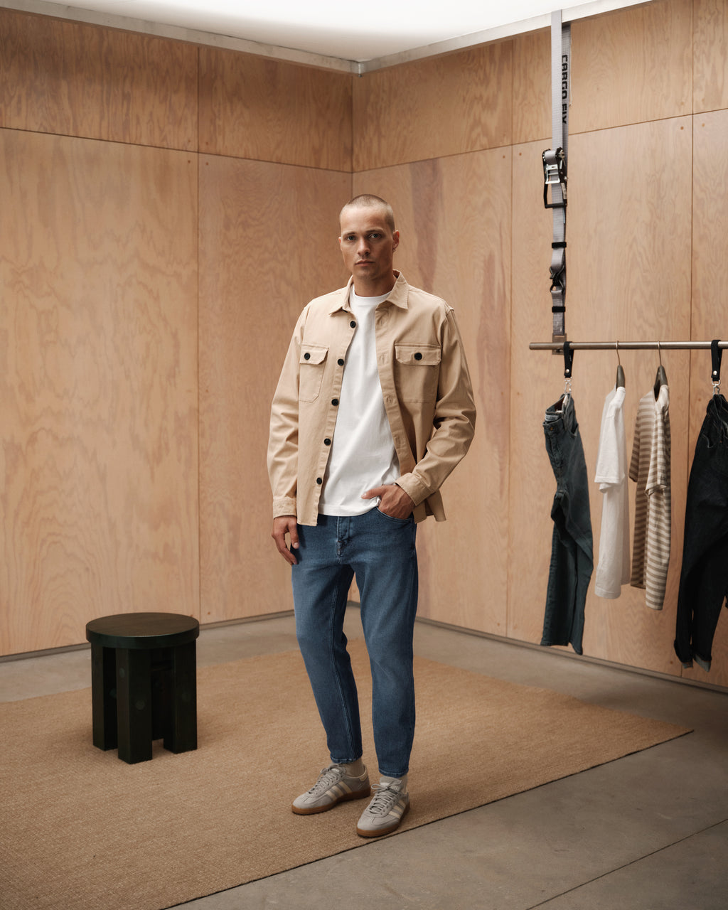 A man stands in a minimalist room with wood-paneled walls, wearing GABBA Alex K3948 Jeans in denim wash, a beige overshirt, white t-shirt, and sneakers. A rack holds clothes nearby; a small dark stool sits to the left.