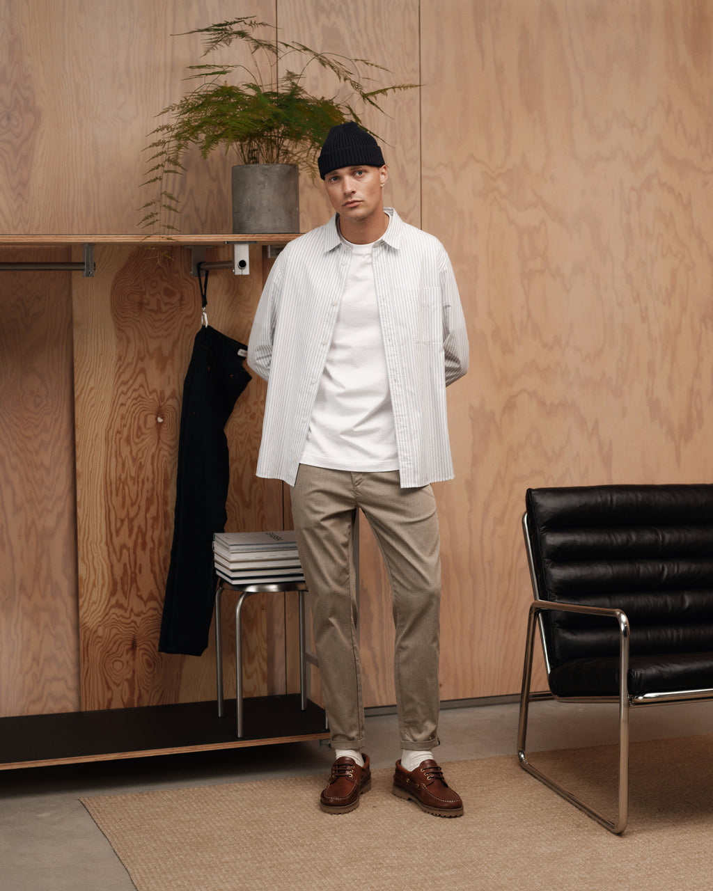 A man in the Gabba Sven Reg Clo LS - White Stripe shirt, black beanie, white t-shirt, beige pants, and brown shoes stands in a modern room with wooden walls beside a chair, stools, and a potted plant.