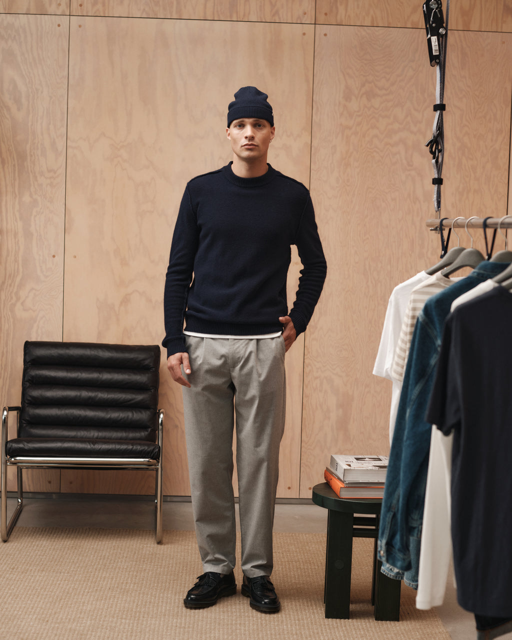 A man wearing a navy sweater, Gabba Napoli 19 Carre pants in Lt. Grey, and a navy beanie stands indoors beside a clothing rack; behind him are a black chair, a wooden wall, and a small table stacked with books.