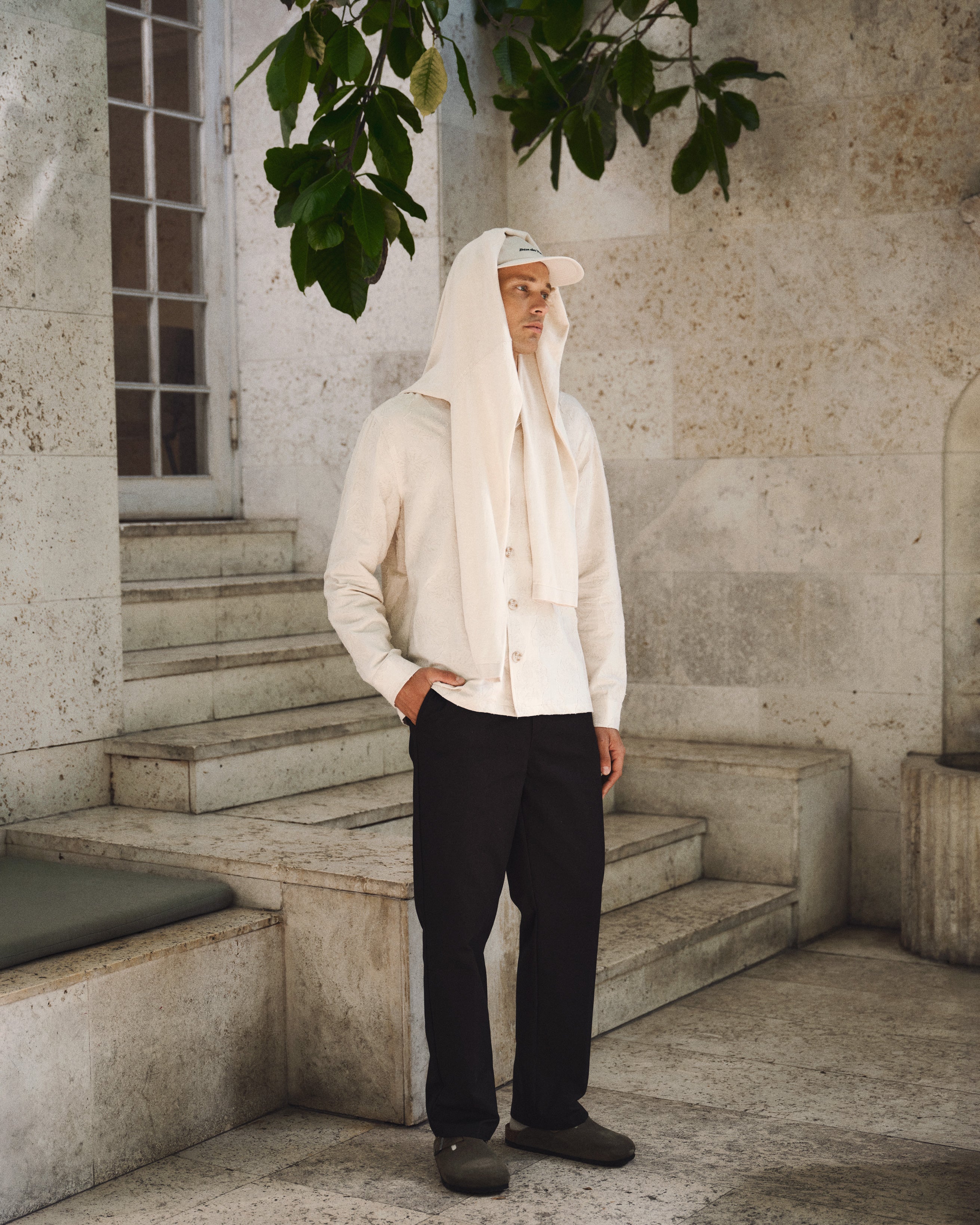 A man stands on stone steps by a wall, wearing the Gabba GABaustin Reso Raw LS Overshirt in Lt. Sand, dark pants, clogs, and a cap with a sweater draped over his head. A leafy branch hangs above as he looks to the side with hands in his pockets.