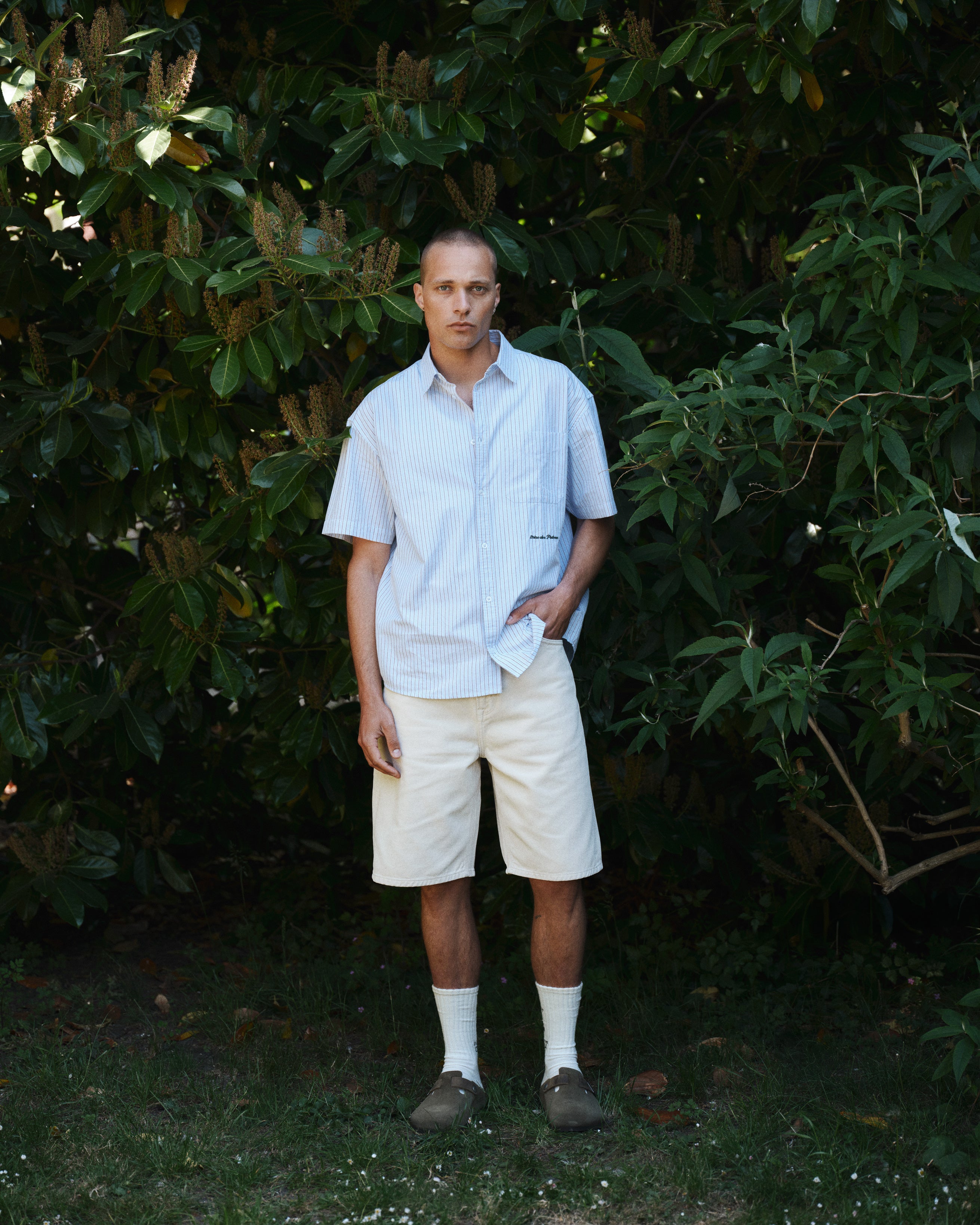 A man stands in front of green foliage, wearing a light blue short-sleeve shirt and Gabba GABmox Antiq Shorts in Ecru, paired with white socks and brown shoes. One hand is in his pocket and he has a neutral expression.