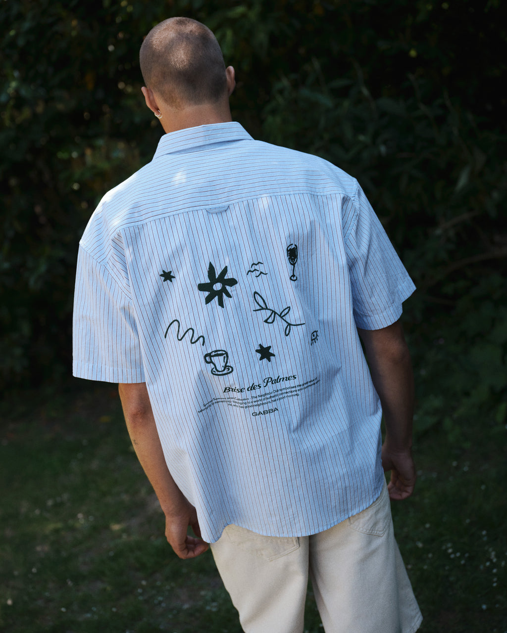 A person stands outdoors with green foliage behind, wearing the Gabba GABjens Over Bisi SS Shirt in brown stripe—a relaxed fit cotton shirt featuring abstract black designs and text on the back—paired with light shorts.