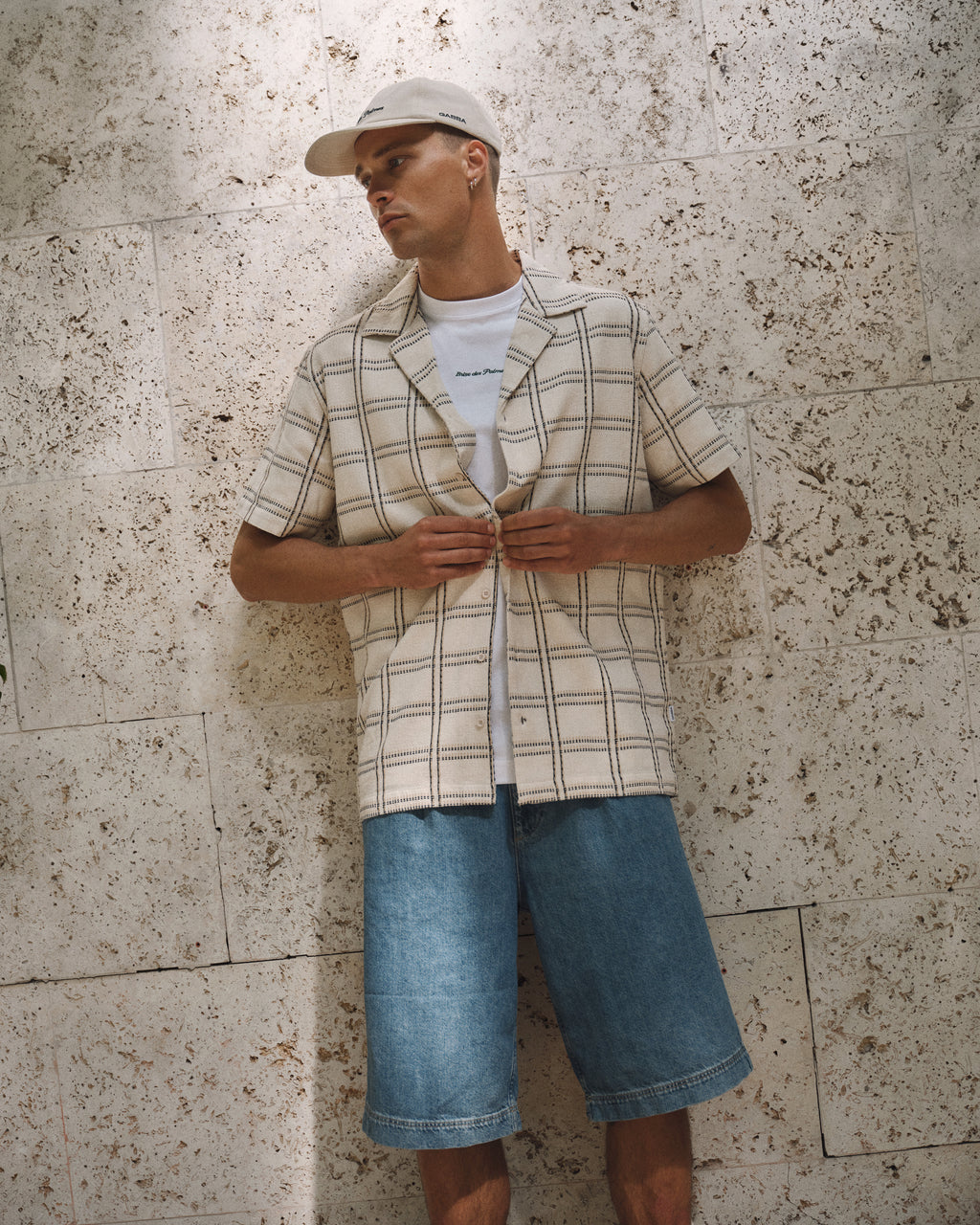 A man wearing the Gabba GABsven Reg Resort Wire SS Shirt in Beige Check stands against a textured stone wall, layered over a white T-shirt and paired with knee-length denim shorts and a beige cap, looking to the side in a relaxed pose.