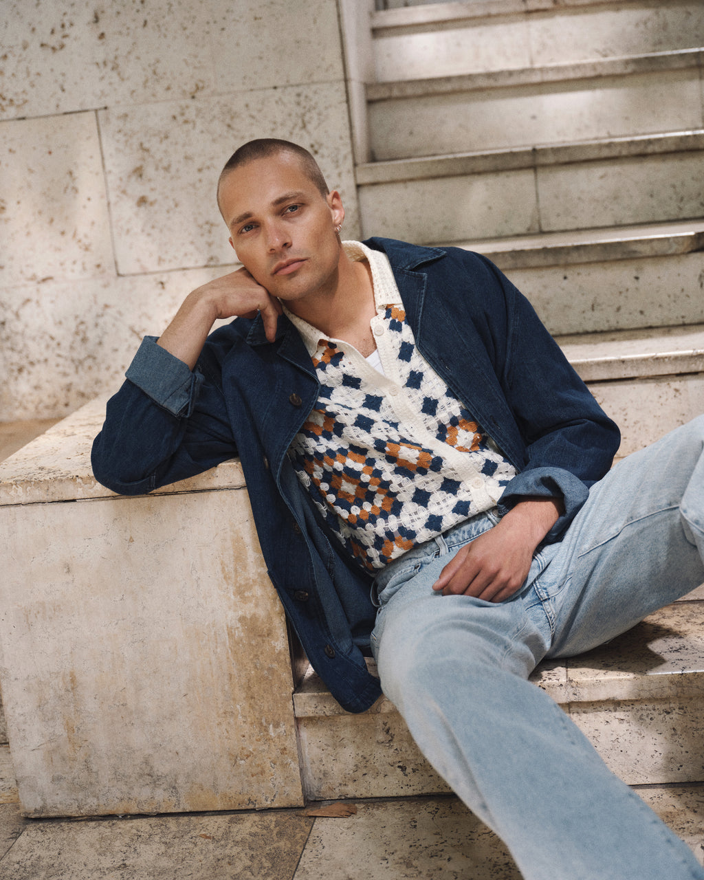 A person with short hair sits on stone steps, wearing a blue jacket, light jeans, and the Gabba GABcrochet Pattern SS Knit - Multi Pattern sweater vest as a layering piece, looking relaxed with one hand resting on their face.