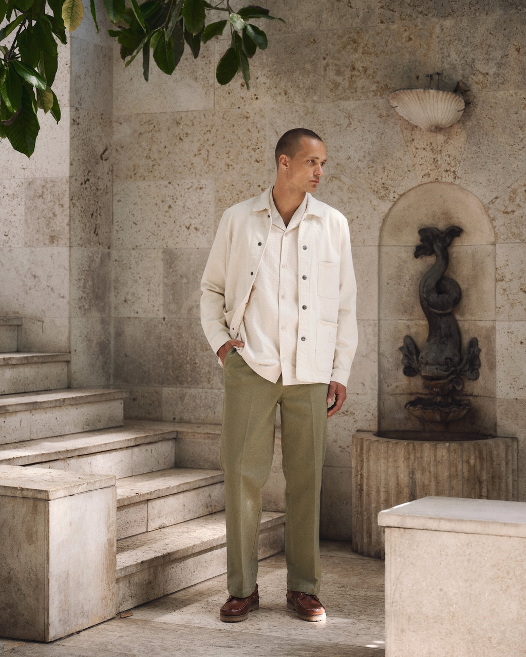 A man stands on stone steps outdoors, wearing Gabba's GABbeta Twill - Ecru overshirt over a white shirt, olive green pants, and brown shoes. Behind him is a stone wall with a decorative fountain and leafy branches overhead.