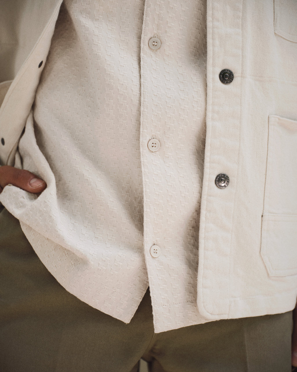 A close-up of a person wearing the Gabba GABtimo Resort Neal SS Shirt in Birch under a cream jacket with metal buttons and a large pocket, paired with olive green pants. Their thumb is hooked into their pocket.