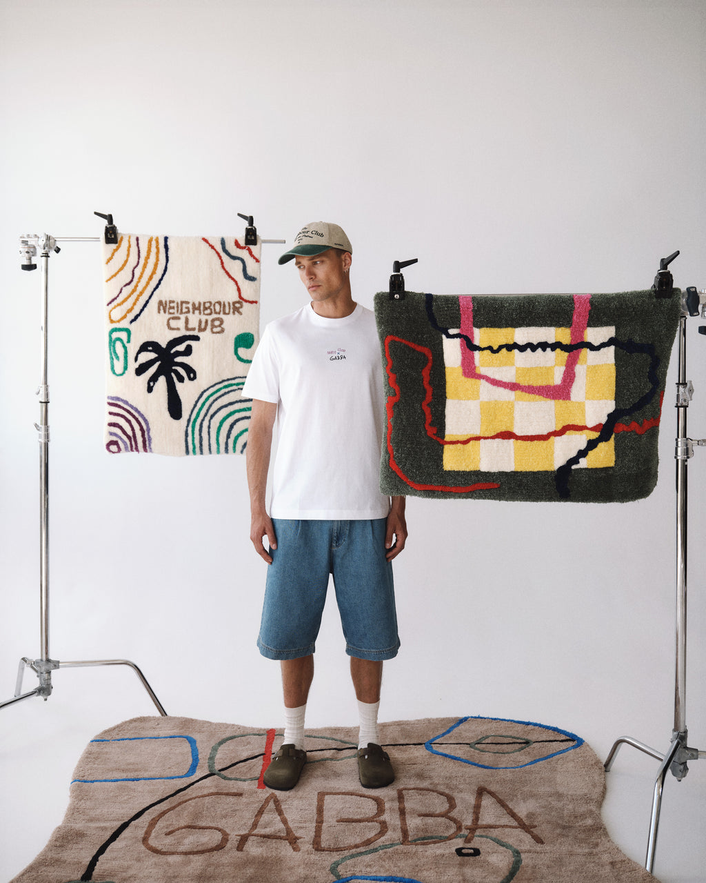A man in a Gabba GABnigel Nando SS T-shirt GOTS - White, denim shorts, cap, and slippers stands between two colorful rugs on stands, with another patterned rug labeled Gabba on the floor, all against a plain white background.