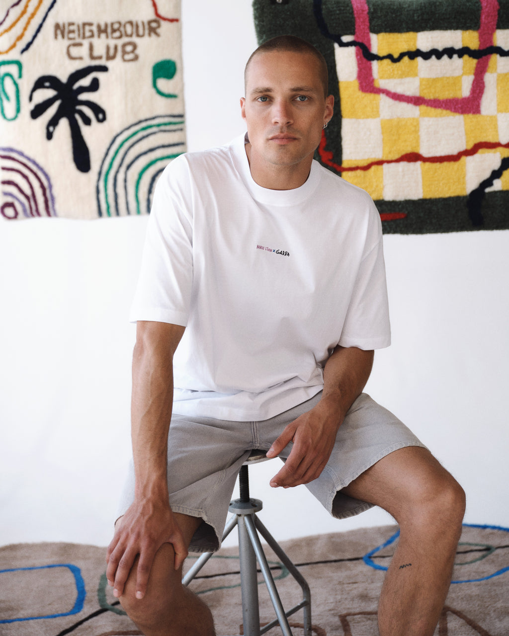 Wearing a Gabba GABnigel Boxy Nando SS T-Shirt GOTS in white and light grey shorts, a person sits on a stool by colorful abstract rugs, with a NEIGHBOUR CLUB sign hanging behind them, creating an artistic, relaxed vibe.