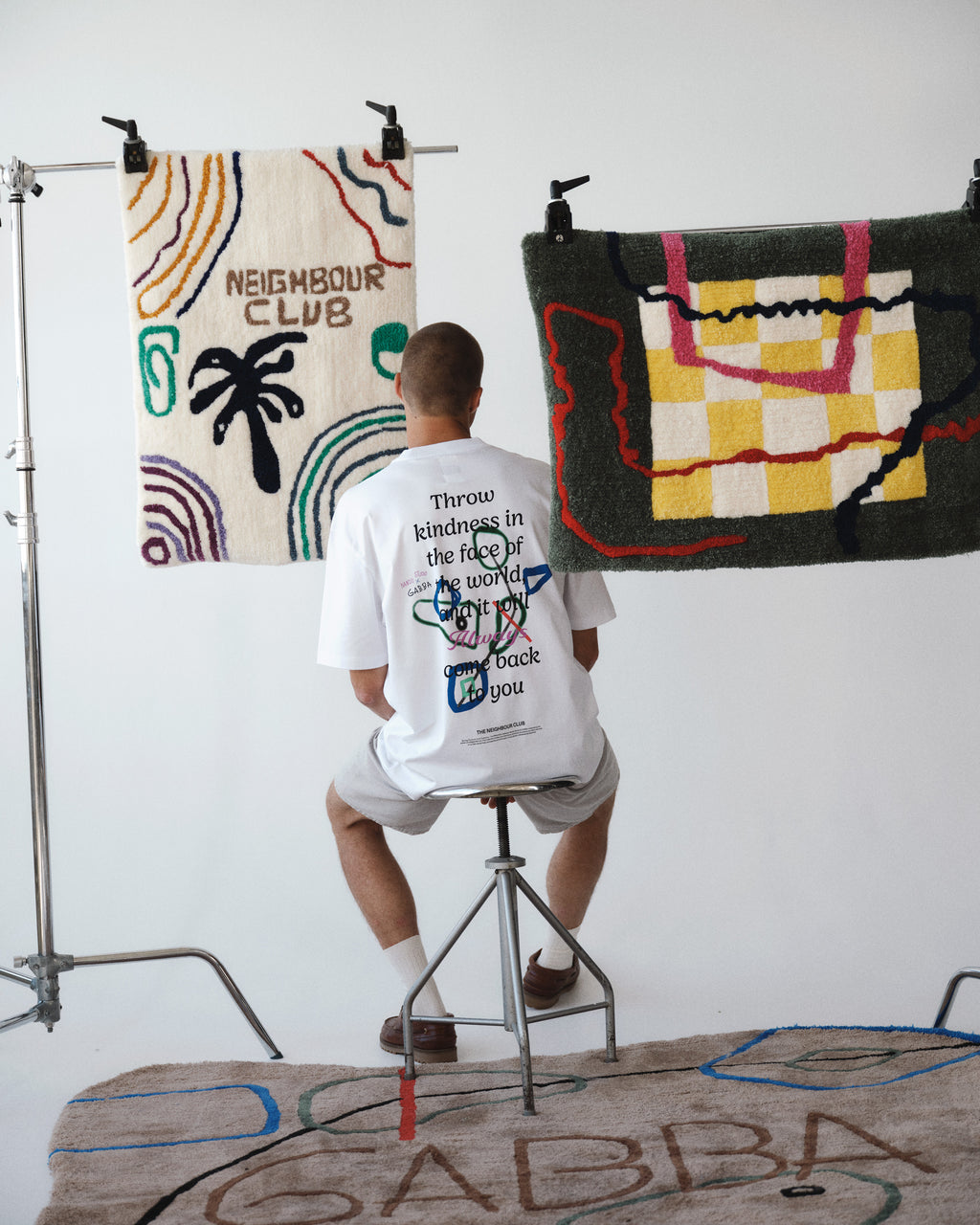 A person sits on a stool wearing the Gabba GABnigel Boxy Nando SS T-Shirt GOTS in white. They face away in a bright studio with abstract rugs behind them and are seated on a large rug that has 